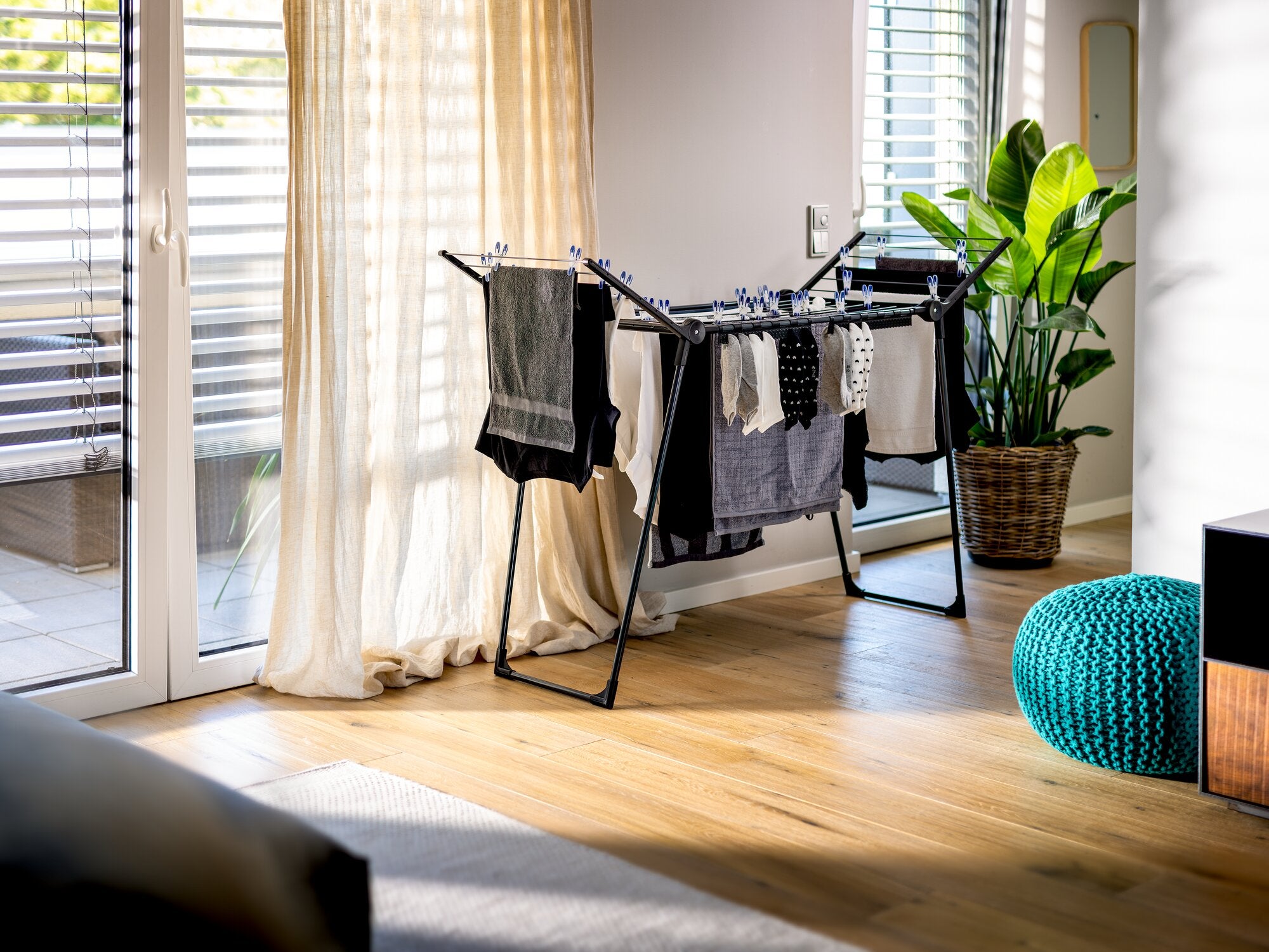 Leifheit Pegasus Slim 15m Black Wäscheständer in einem hellen Wohnraum am Fenster, aufgeklappt mit dunkler und heller Wäsche behängt, auf Holzfußboden neben Zimmerpflanze und blauem Sitzkissen.