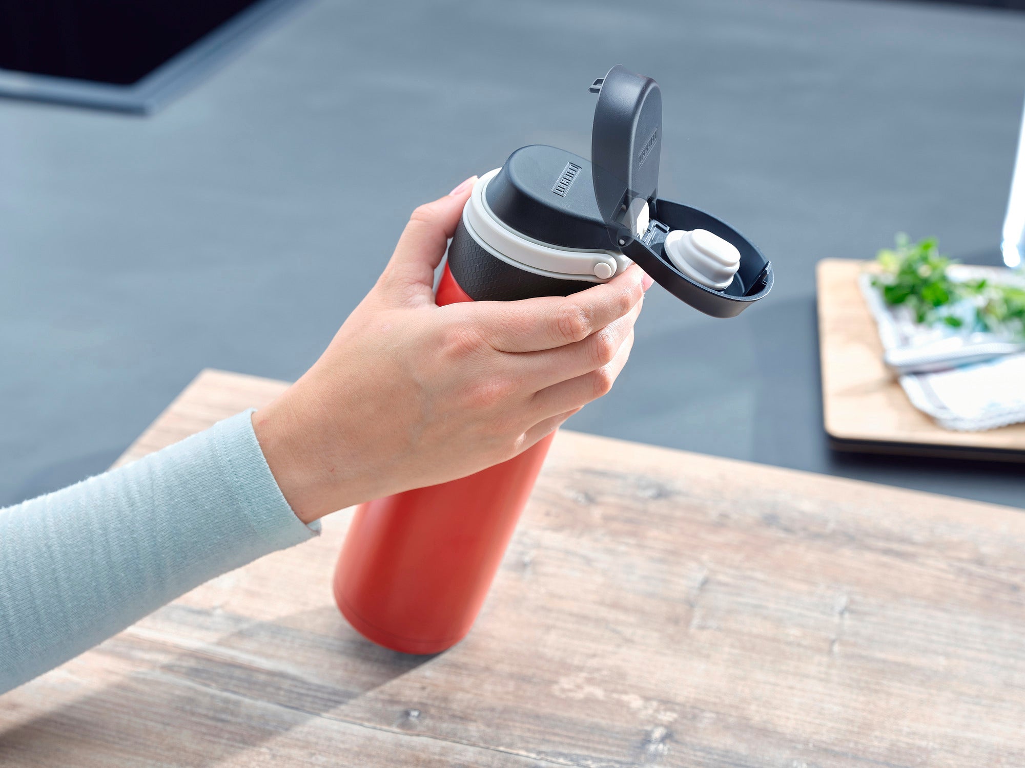 Isolierbecher Flip 600ml in Rot wird auf einem Tisch verwendet. Eine Hand öffnet den Deckel, um den Becher zu benutzen. Im Hintergrund sind Küchenutensilien zu sehen.