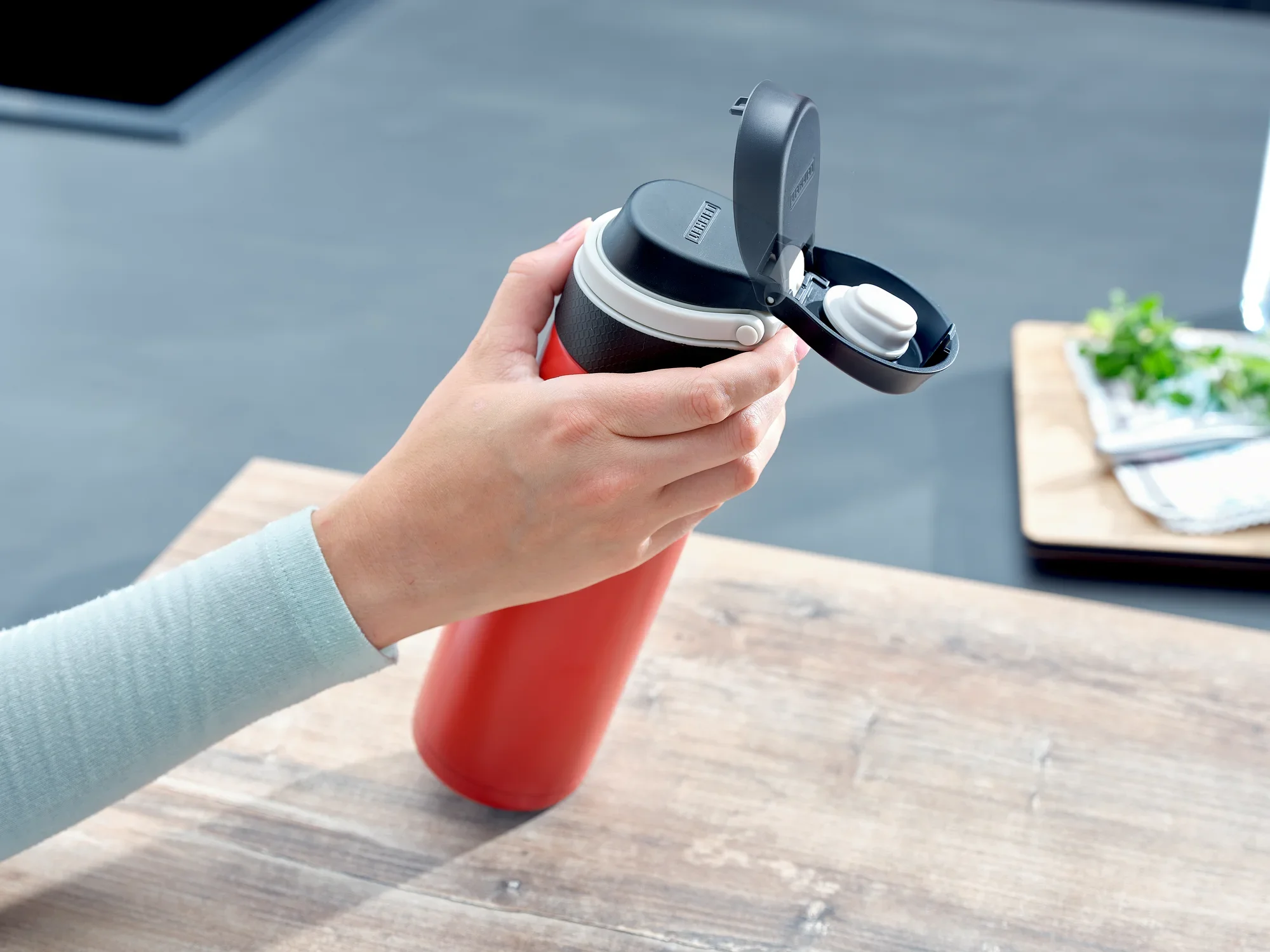 Izolowany kubek Flip 600 ml w kolorze czerwonym jest używany na stole. Ręka otwiera pokrywkę, aby użyć kubka. W tle widoczne są przybory kuchenne.