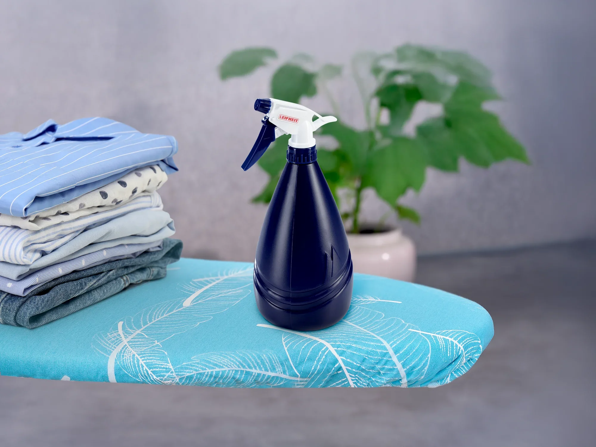 Un vaporisateur bleu foncé est posé sur une table à repasser à côté d'une pile de vêtements pliés. Une plante verte est visible en arrière-plan. Idéal pour le repassage.