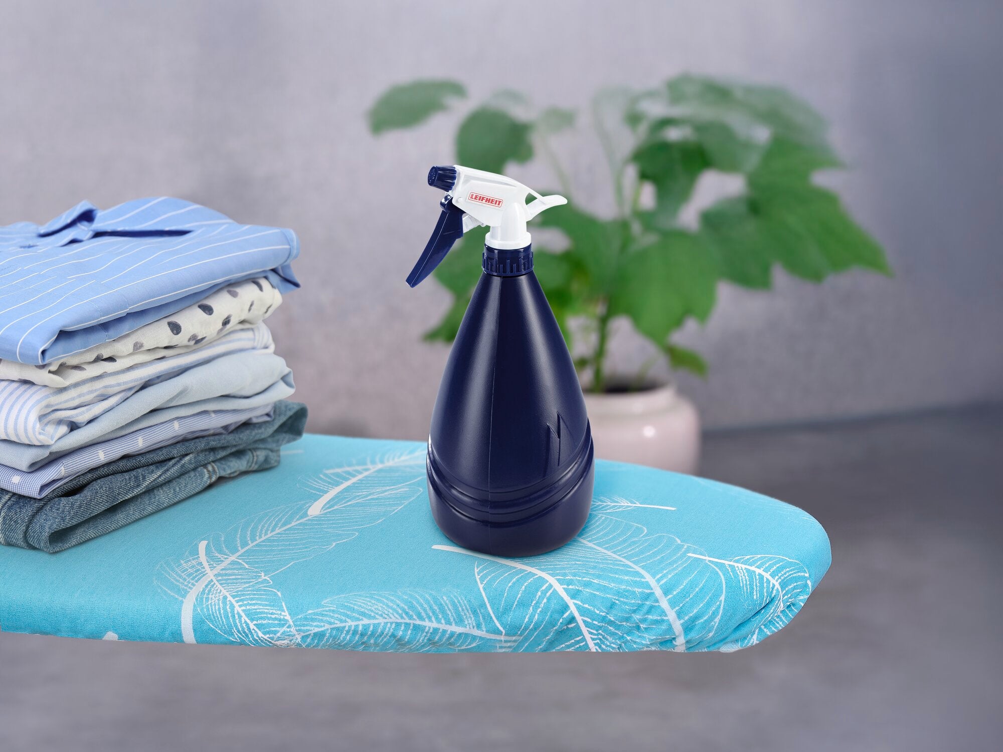 Un vaporisateur bleu foncé est posé sur une table à repasser à côté d'une pile de vêtements pliés. Une plante verte est visible en arrière-plan. Idéal pour le repassage.