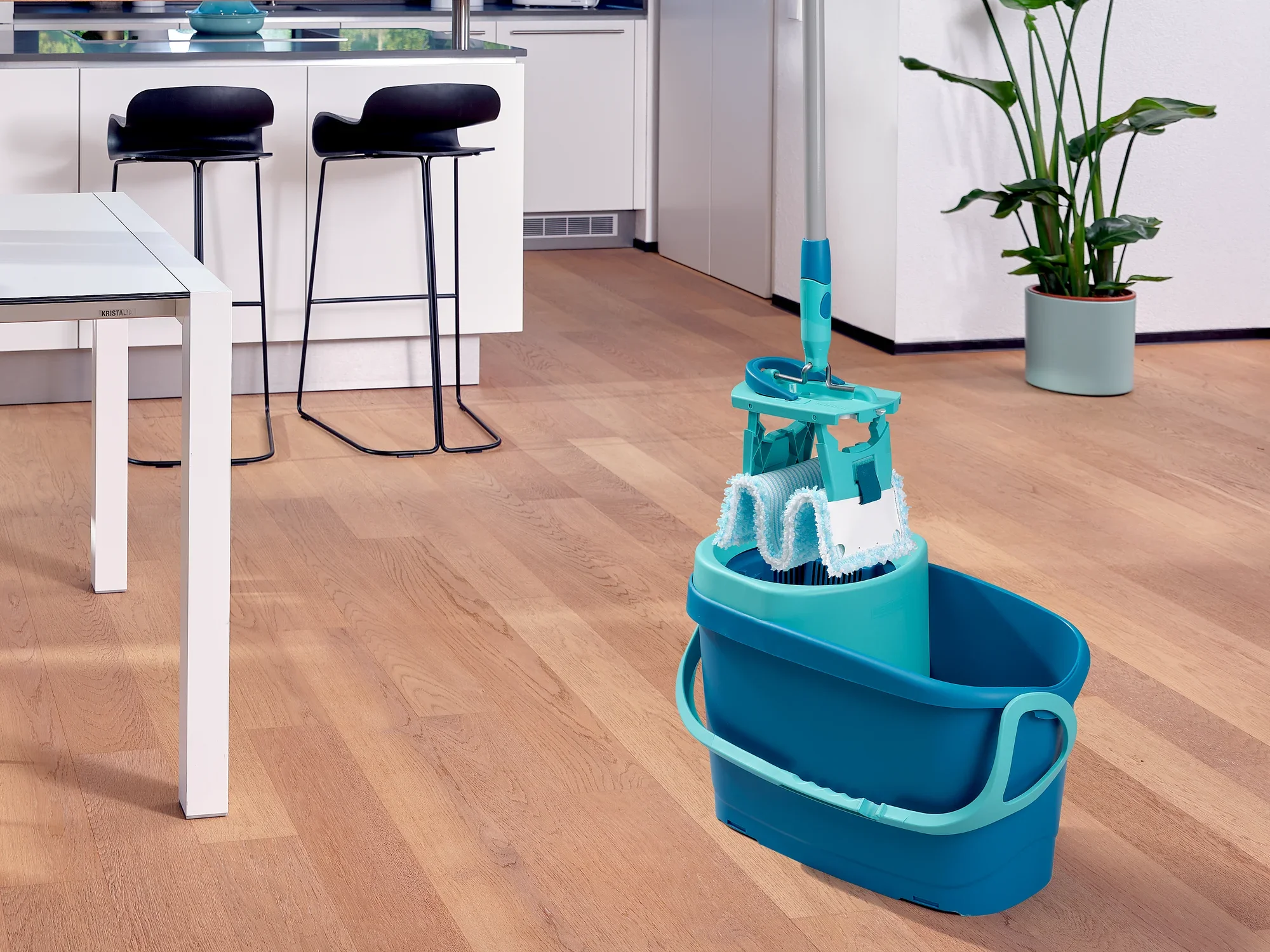 Un set CLEAN TWIST M Ergo se encuentra en una cocina moderna con suelo de madera. El cubo es de color turquesa y contiene una mopa a juego. Al fondo se ven taburetes de bar y una planta.
