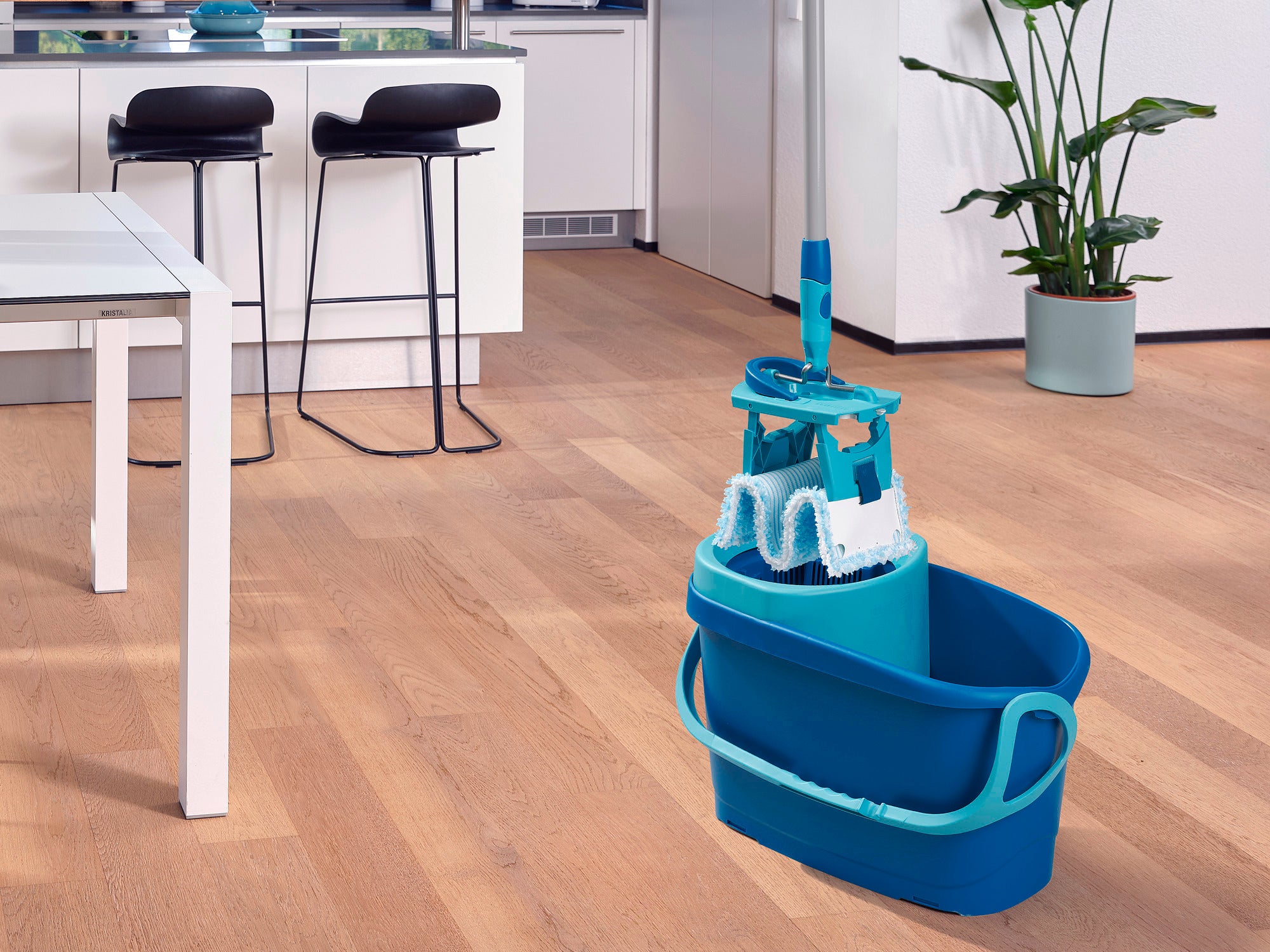 Een CLEAN TWIST M Ergo Set is geplaatst in een moderne keuken met een houten vloer. De emmer is turkoois en bevat een bijpassende mop. Op de achtergrond staan barkrukken en een plant.