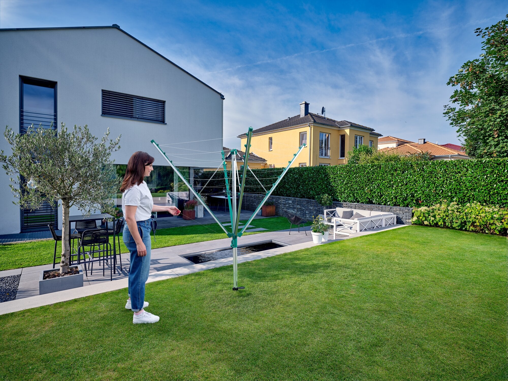 Eine Frau steht in einem gepflegten Garten und benutzt die Wäschespinne Linomatic V 400. Im Hintergrund ist ein modernes Haus mit Terrasse und Gartenmöbeln zu sehen.