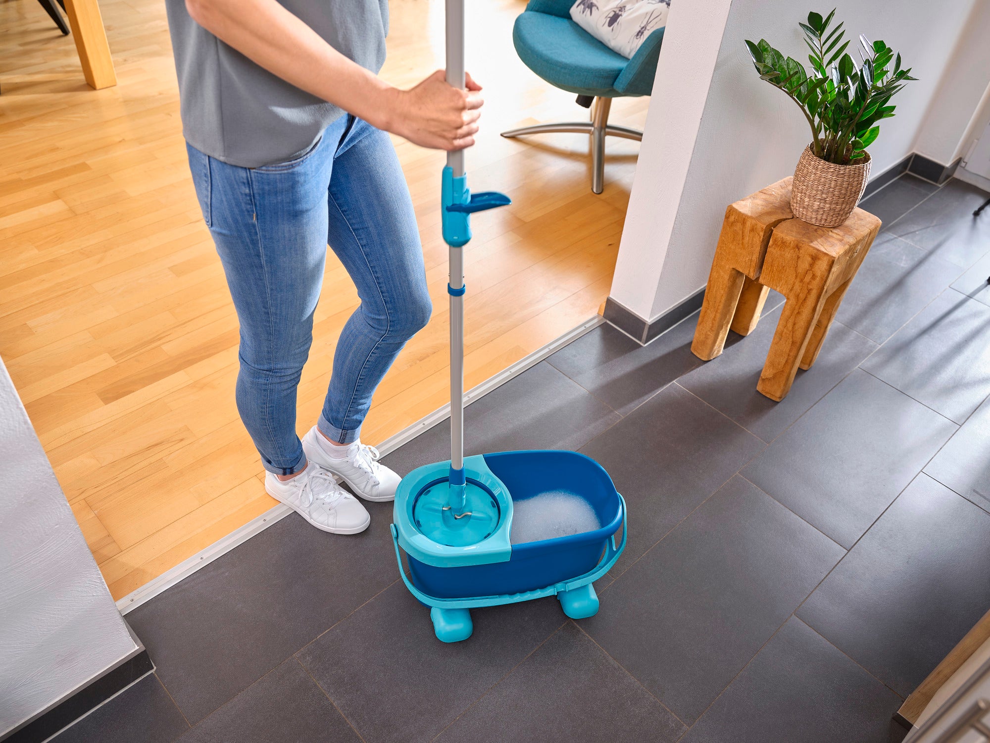 Eine Person verwendet den CLEAN TWIST Disc Mop Ergo in einem modernen Wohnzimmer. Der Mop ist in einem Eimer mit Wasser, bereit für die Reinigung von Fliesenböden.