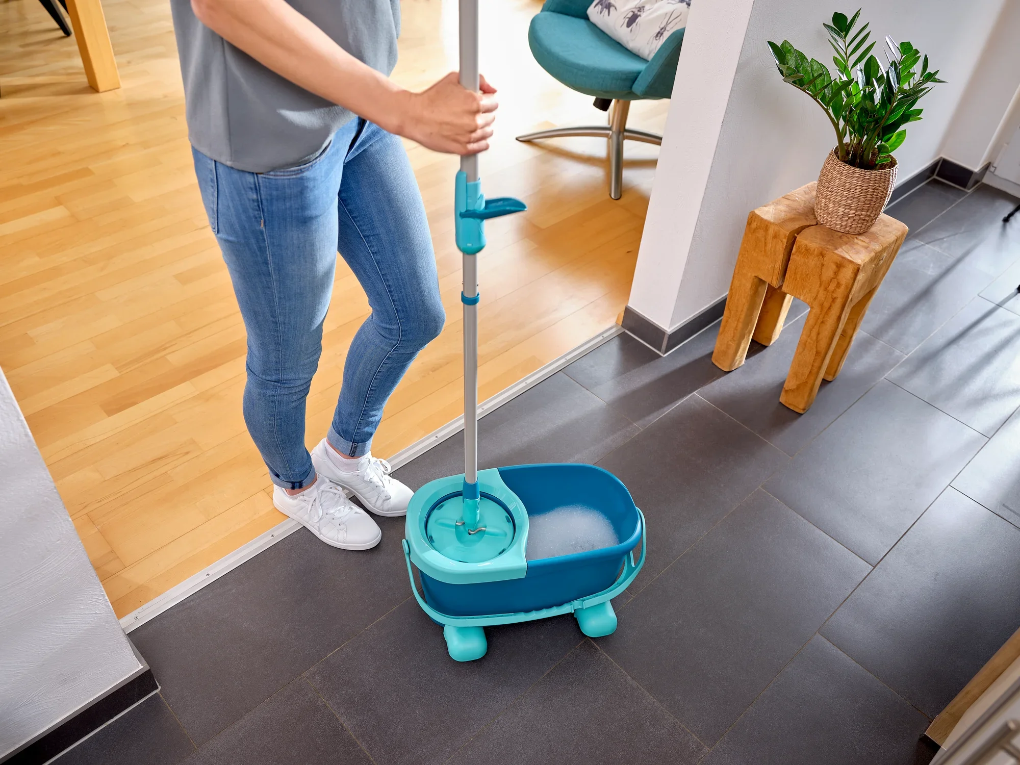 Een persoon gebruikt de CLEAN TWIST Disc Mop Ergo in een moderne woonkamer. De mop ligt in een emmer water, klaar om tegelvloeren schoon te maken.