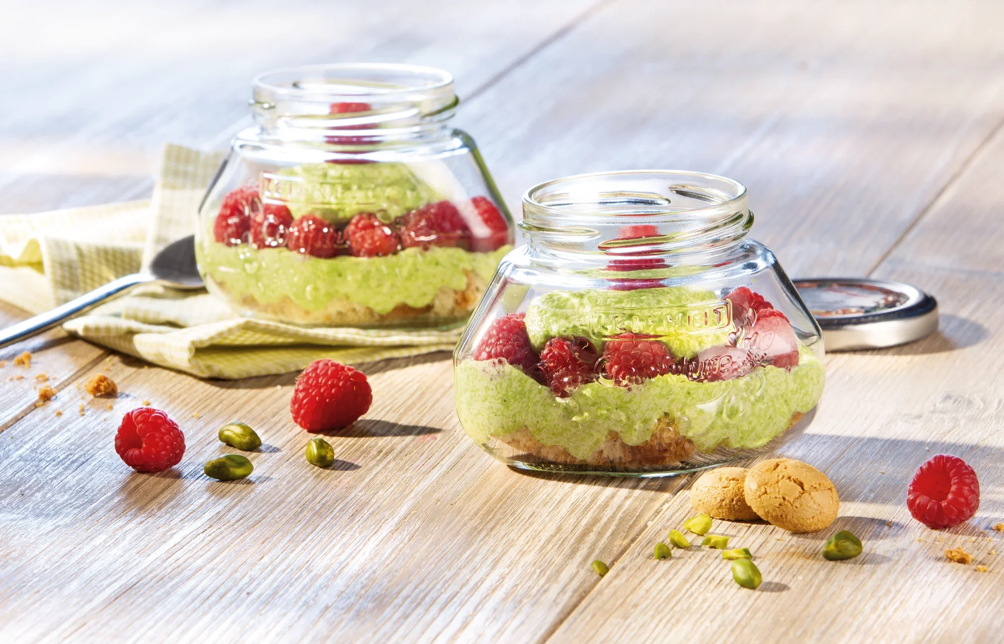 Dwa słoiki o pojemności 250 ml stoją na drewnianym stole. Wypełnione są zielonym kremem, malinami i herbatnikami. W tle rozsypane są pistacje i herbatniki.