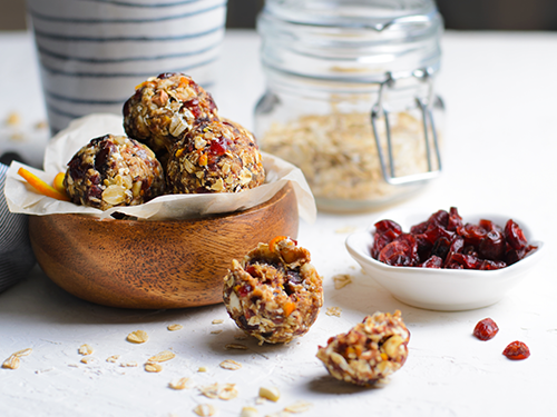 Eine Holzschale mit Energy-Balls steht schoen angerichtet neben einer Schale mit getrockneten Cranberries und einem Glas Haferflocken auf einem Tisch.
