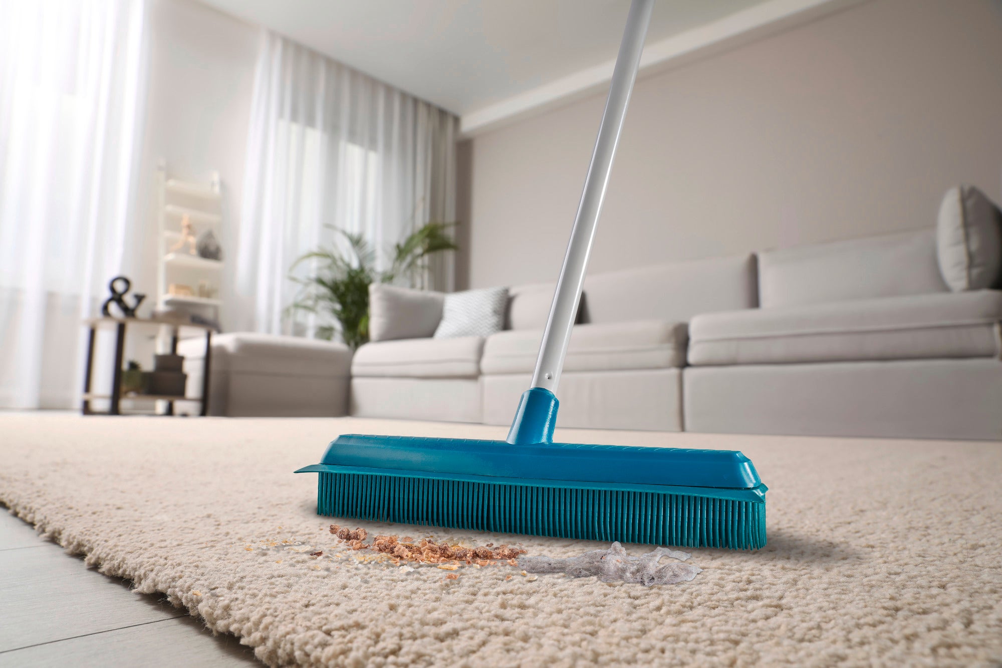 Een turquoise superbezem reinigt een lichtgekleurd tapijt in een moderne woonkamer. De bezem verwijdert effectief vuil en stof, terwijl op de achtergrond een gezellige sofa te zien is.