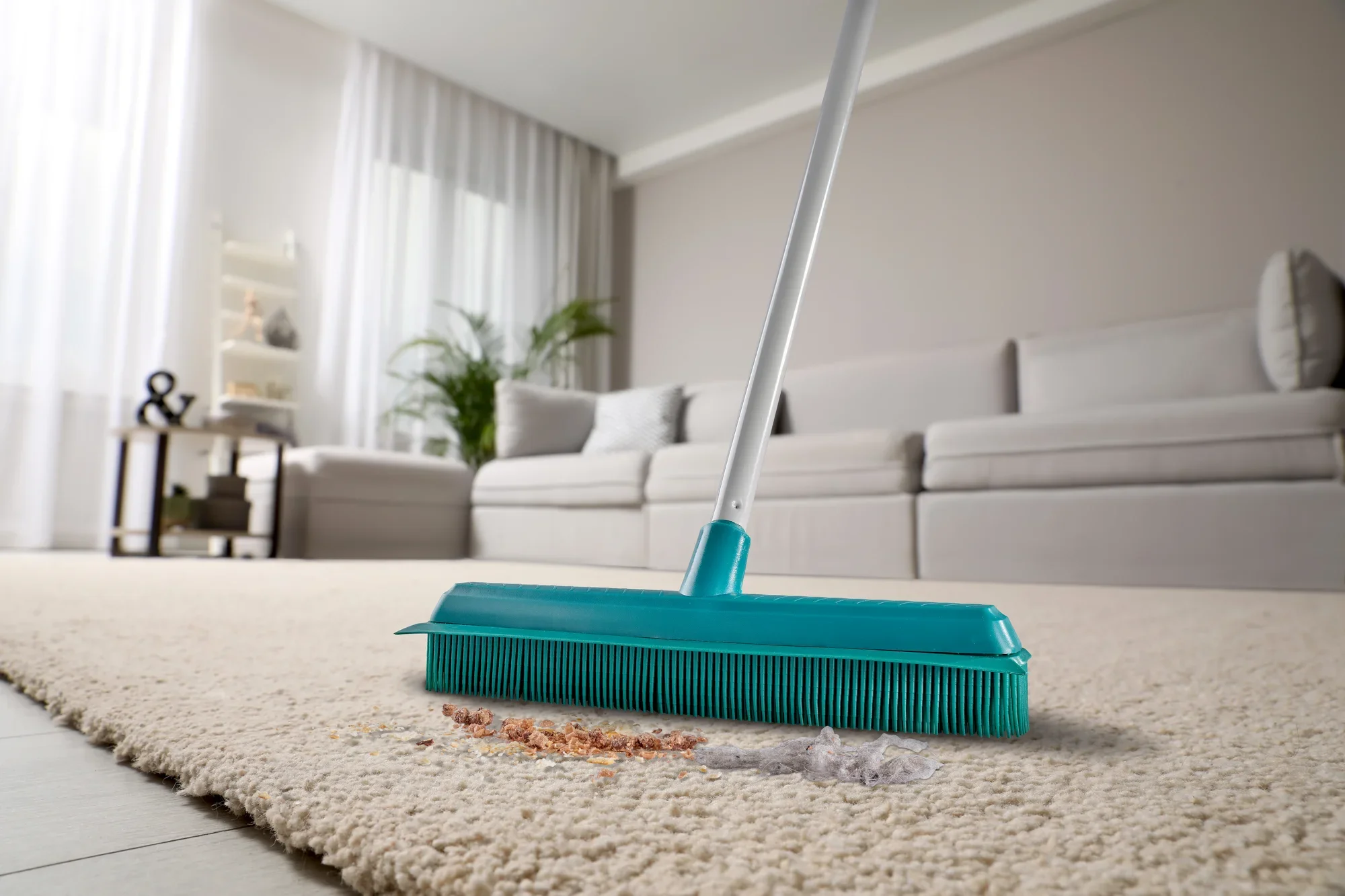 Una scopa Suprabesen turchese pulisce un tappeto chiaro in un salotto moderno. La scopa rimuove efficacemente sporco e polvere, mentre sullo sfondo si vede un comodo divano.