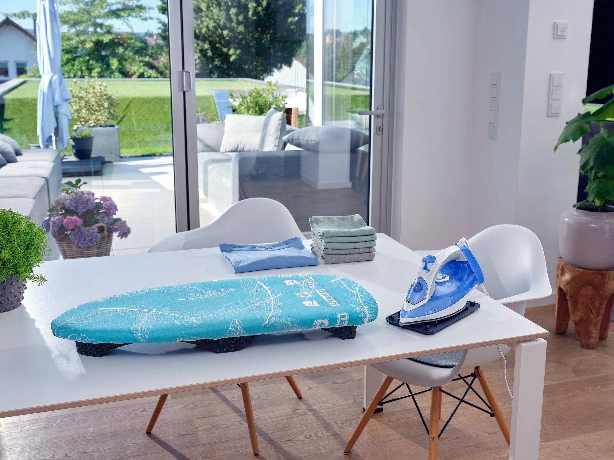 Air Board Table Compact ironing table on a modern dining table in a bright room with a view of a sunny garden. Next to it is a blue iron and folded laundry.