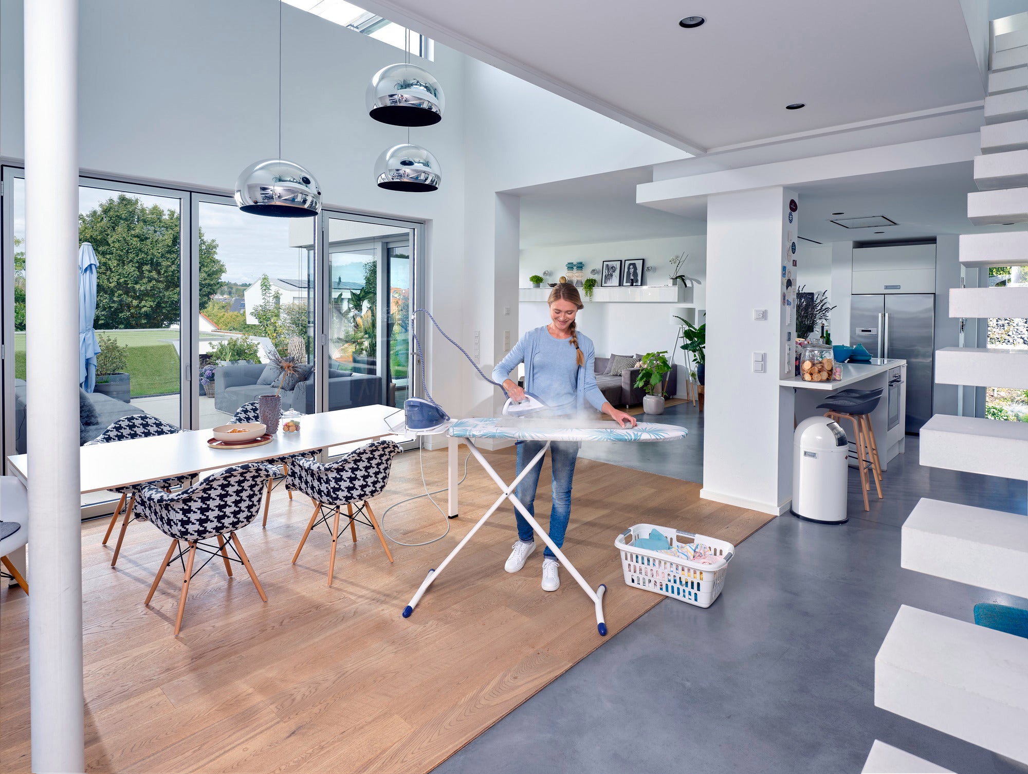 Table à repasser Air Board Express M Solid dans une cuisine moderne. Une personne repasse une chemise tandis que la lumière du soleil pénètre par de grandes fenêtres. La pièce est lumineuse et élégamment meublée.