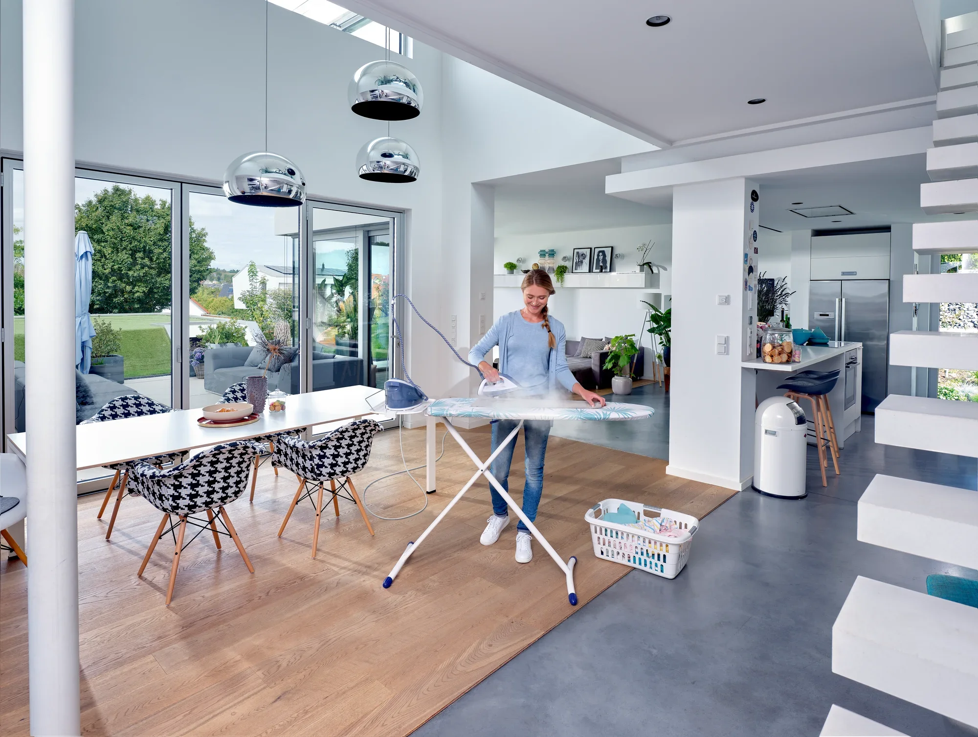 Table à repasser Air Board Express M Solid dans une cuisine moderne. Une personne repasse une chemise tandis que la lumière du soleil pénètre par de grandes fenêtres. La pièce est lumineuse et élégamment meublée.