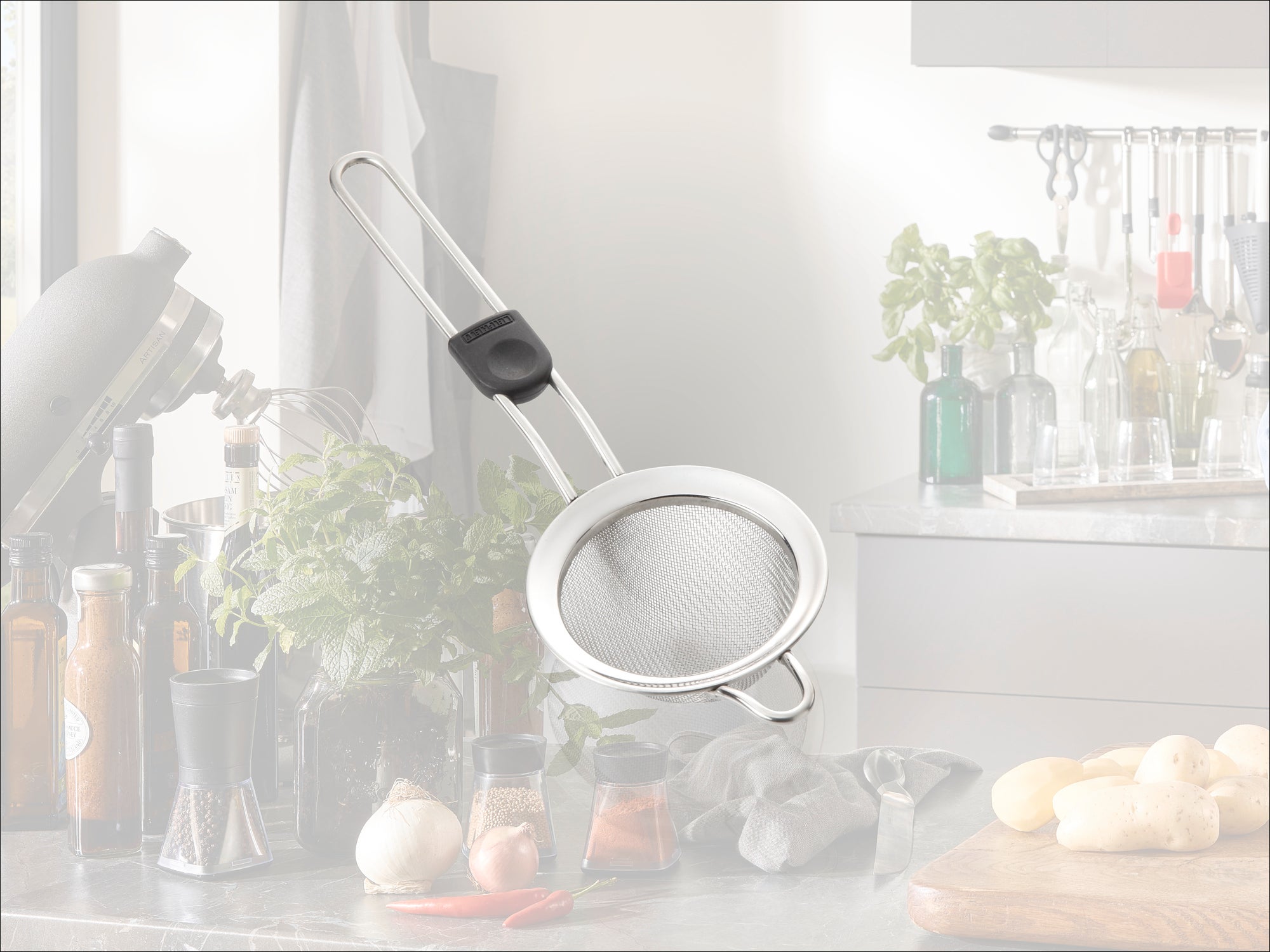 Un colino da cucina con manico lungo e impugnatura nera è appoggiato su un moderno piano di lavoro della cucina. Sullo sfondo si vedono erbe aromatiche fresche e spezie.