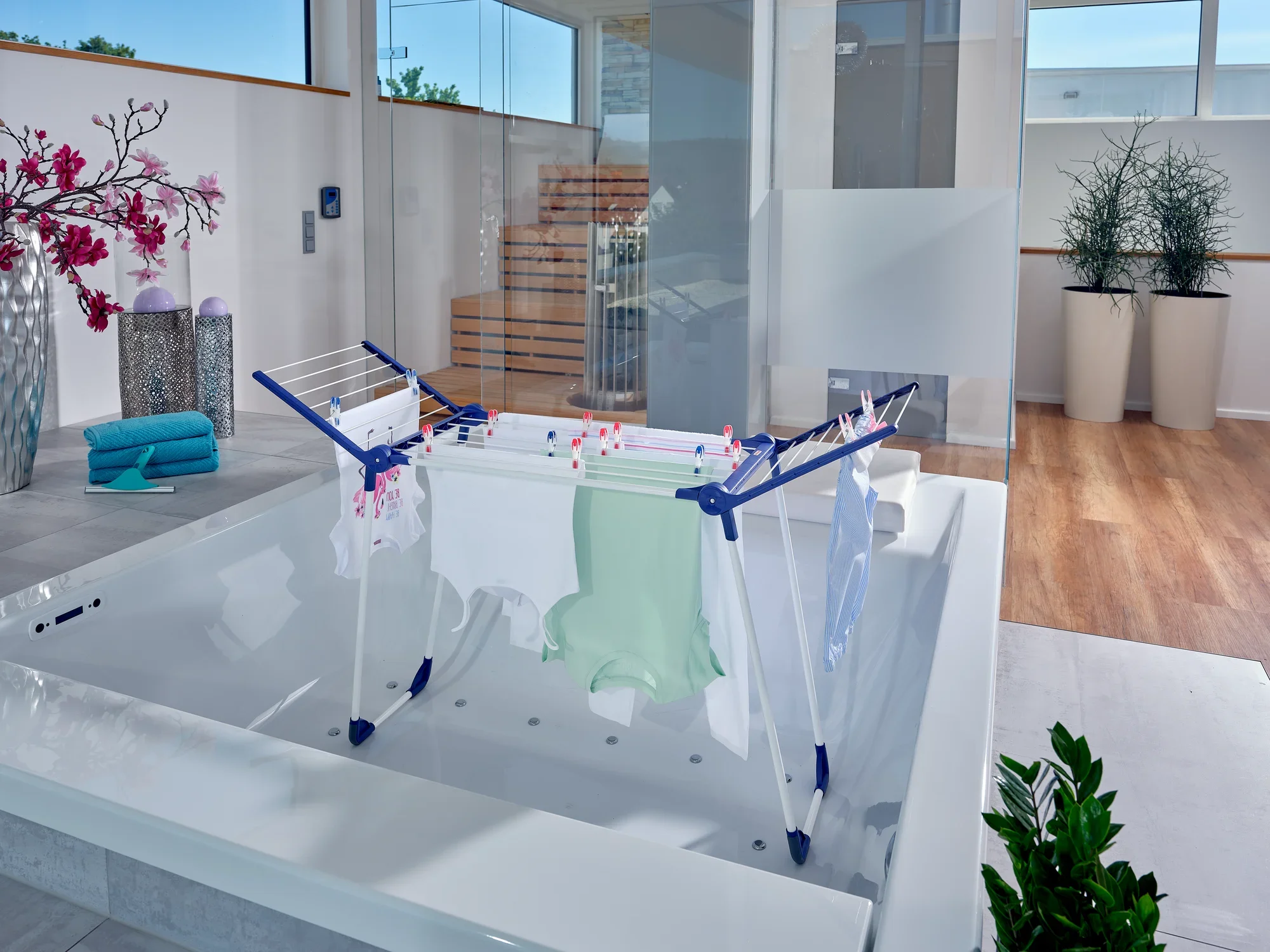 Wolnostojąca suszarka Pegasus Bath stoi na dużej, białej wannie w nowoczesnej łazience. Otoczona roślinami i ręcznikami, delikatnie suszy rozwieszone pranie.