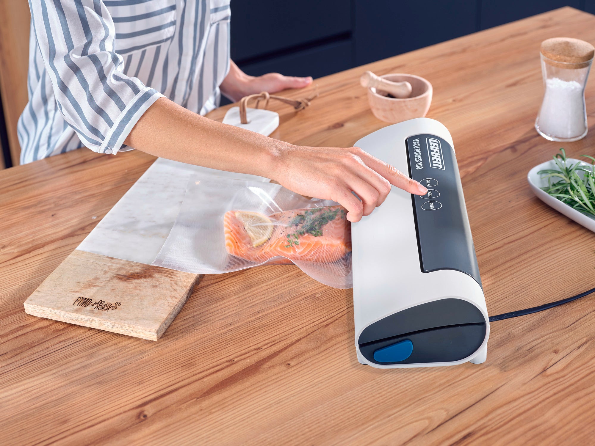 Vakuumiergerät Vacu Power 100 in Aktion: Eine Person bedient das Gerät, um Lachs in einem Beutel zu vakuumieren. Der Tisch ist mit Gewürzen und Kräutern dekoriert.