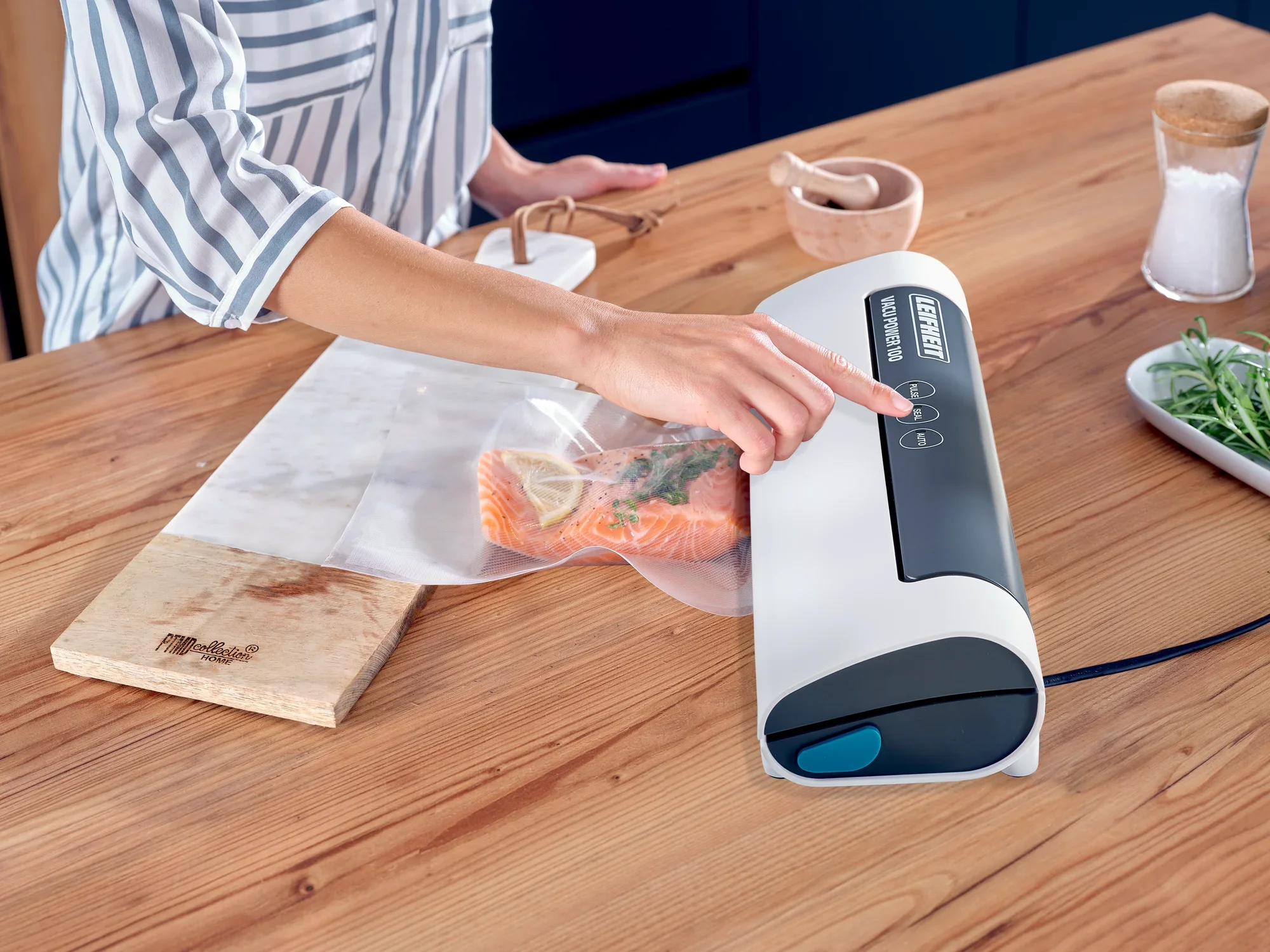 Zgrzewarka próżniowa Vacu Power 100 w akcji: Osoba obsługuje urządzenie w celu próżniowego zamknięcia łososia w woreczku. Stół jest udekorowany przyprawami i ziołami.