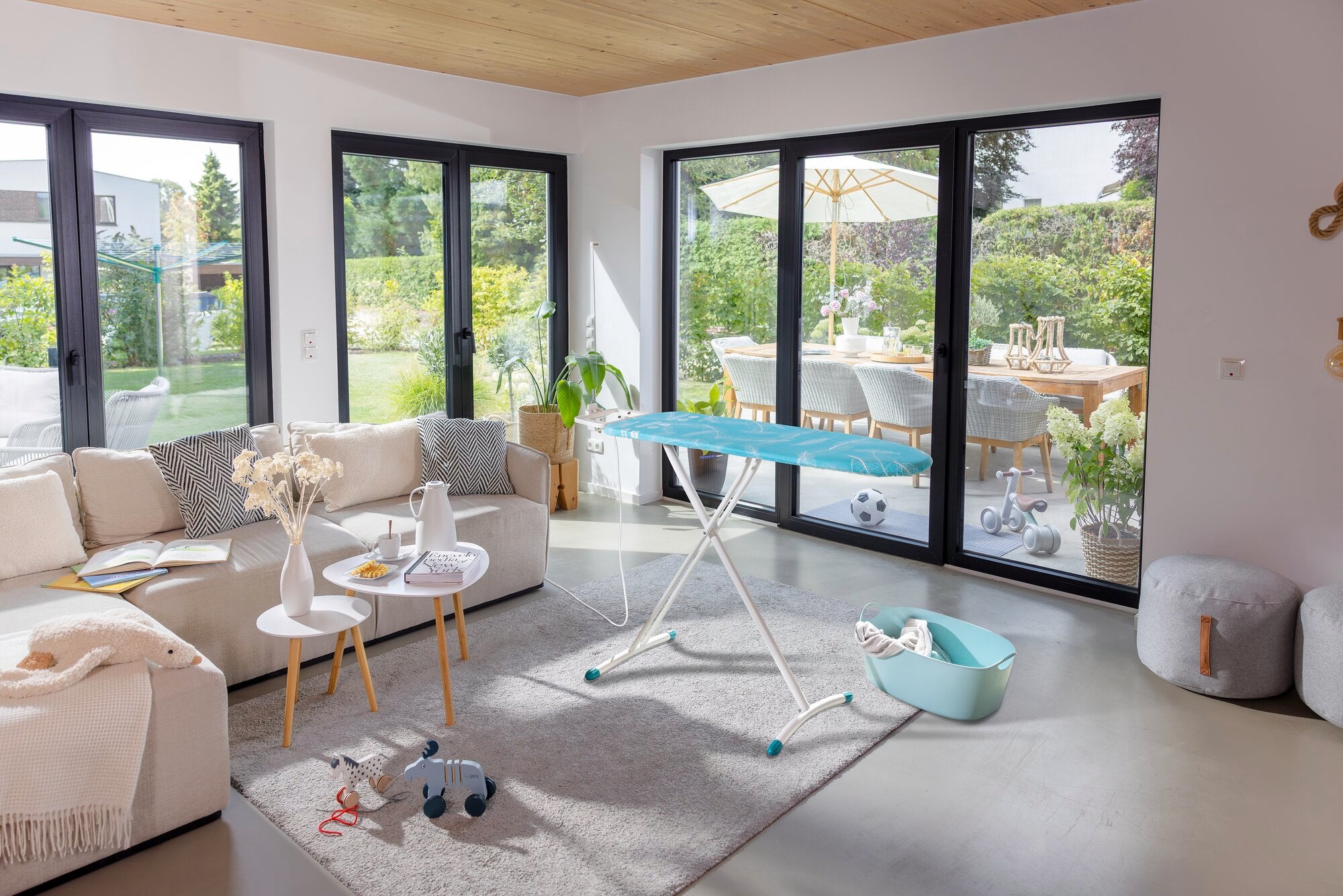 Bügelbrett Leifheit Air Board M Solid Plus in einem modernen Wohnzimmer mit großen Fenstern. Es steht auf einem Teppich, umgeben von einem Sofa und einem Tisch.