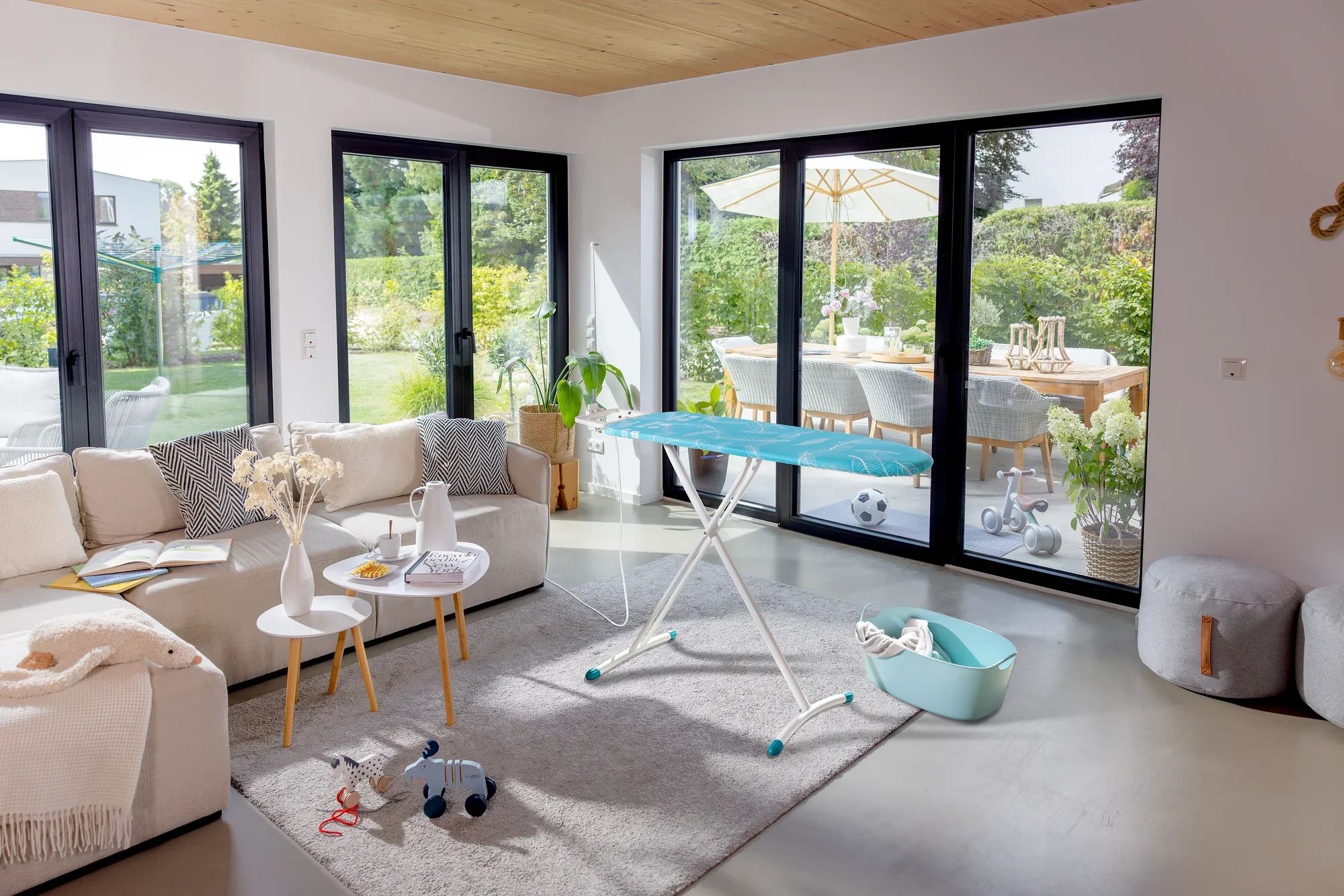 Bügelbrett Leifheit Air Board M Solid Plus in einem modernen Wohnzimmer mit großen Fenstern. Es steht auf einem Teppich, umgeben von einem Sofa und einem Tisch.