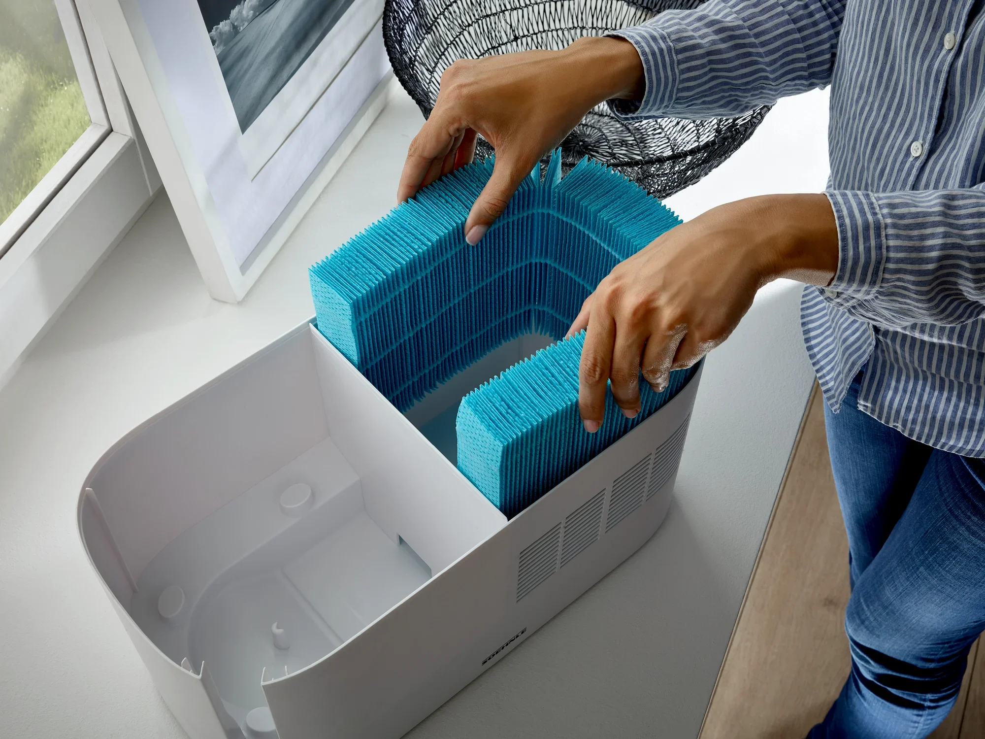 Osoba wkłada niebieski filtr do oczyszczacza powietrza. Oczyszczacz powietrza stoi na parapecie obok obrazu i ozdobnego koszyka. Idealny do zapewnienia czystego powietrza w pomieszczeniu.