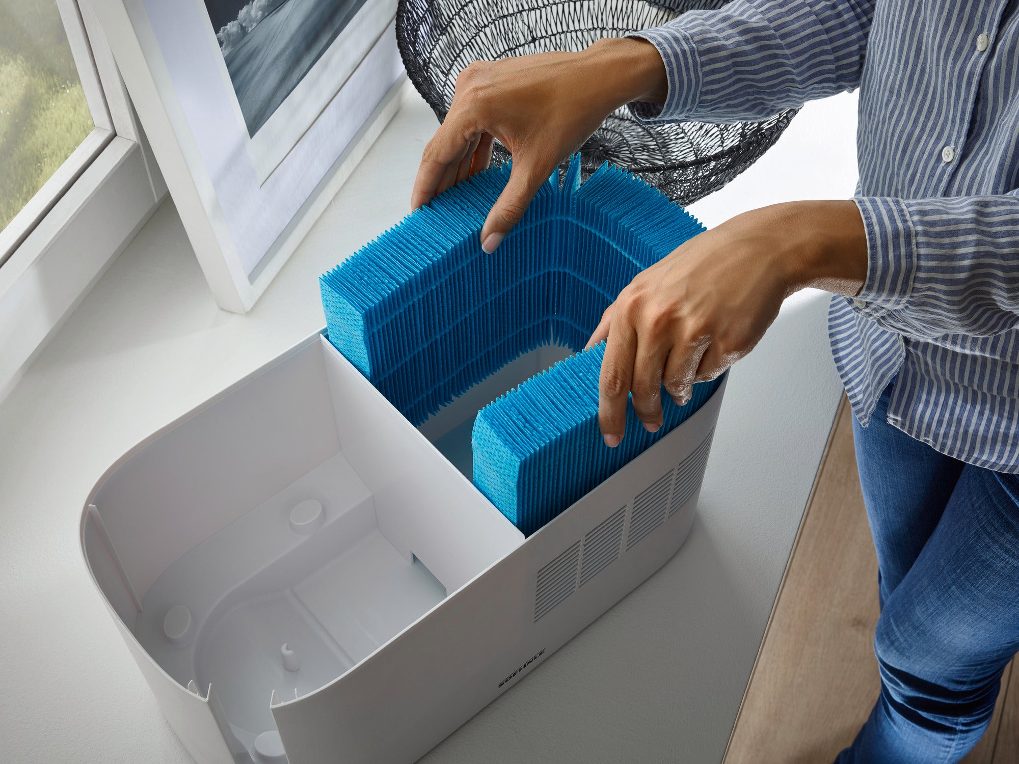 A person inserts a blue filter into an air purifier. The air purifier stands on a windowsill next to a picture and a decorative basket. Ideal for clean air in the room.