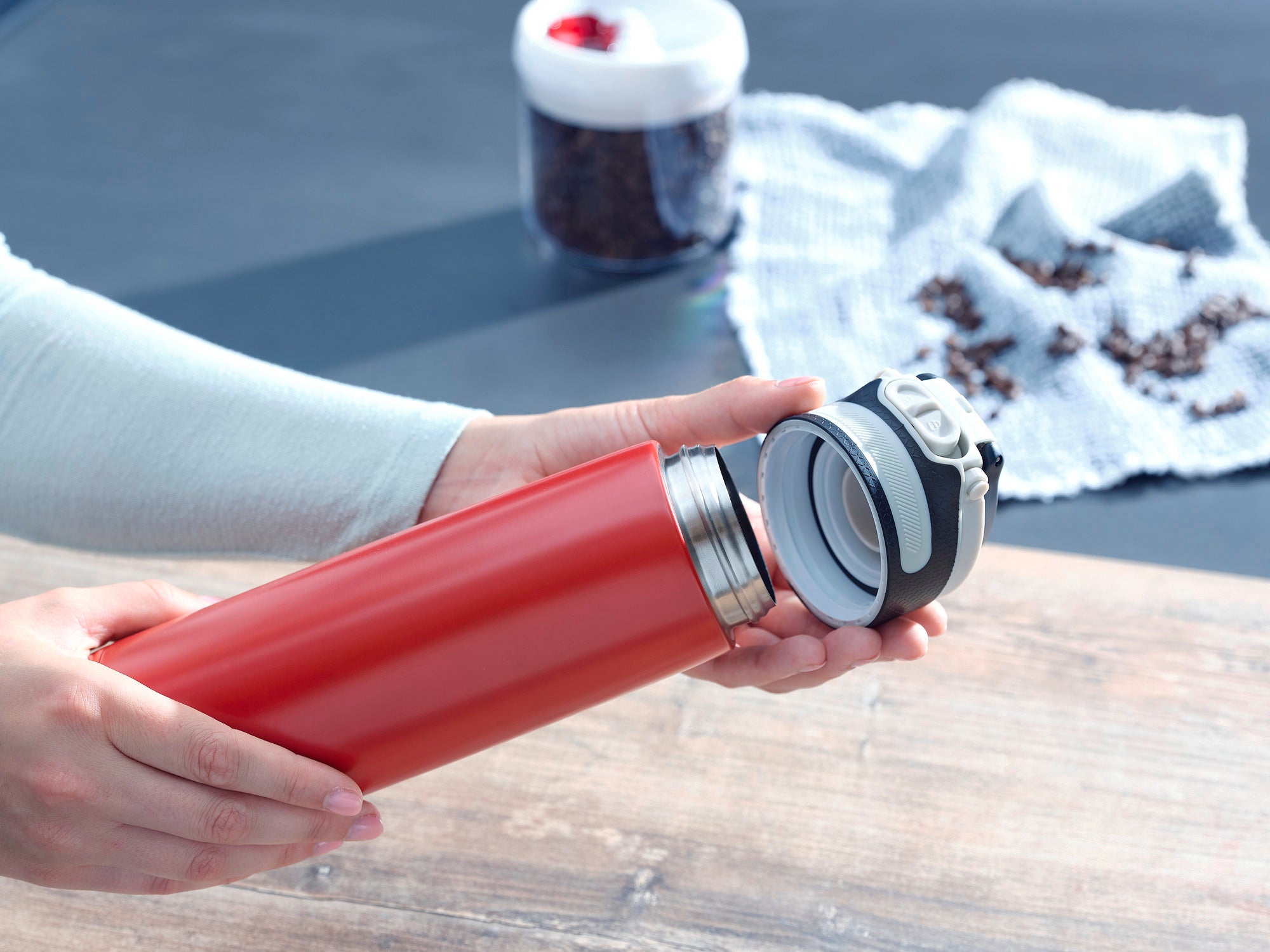 Ein roter Isolierbecher Flip mit 600 ml Fassungsvermögen wird von einer Person geöffnet. Im Hintergrund sind Kaffeebohnen und ein Glasbehälter zu sehen. Ideal für unterwegs.