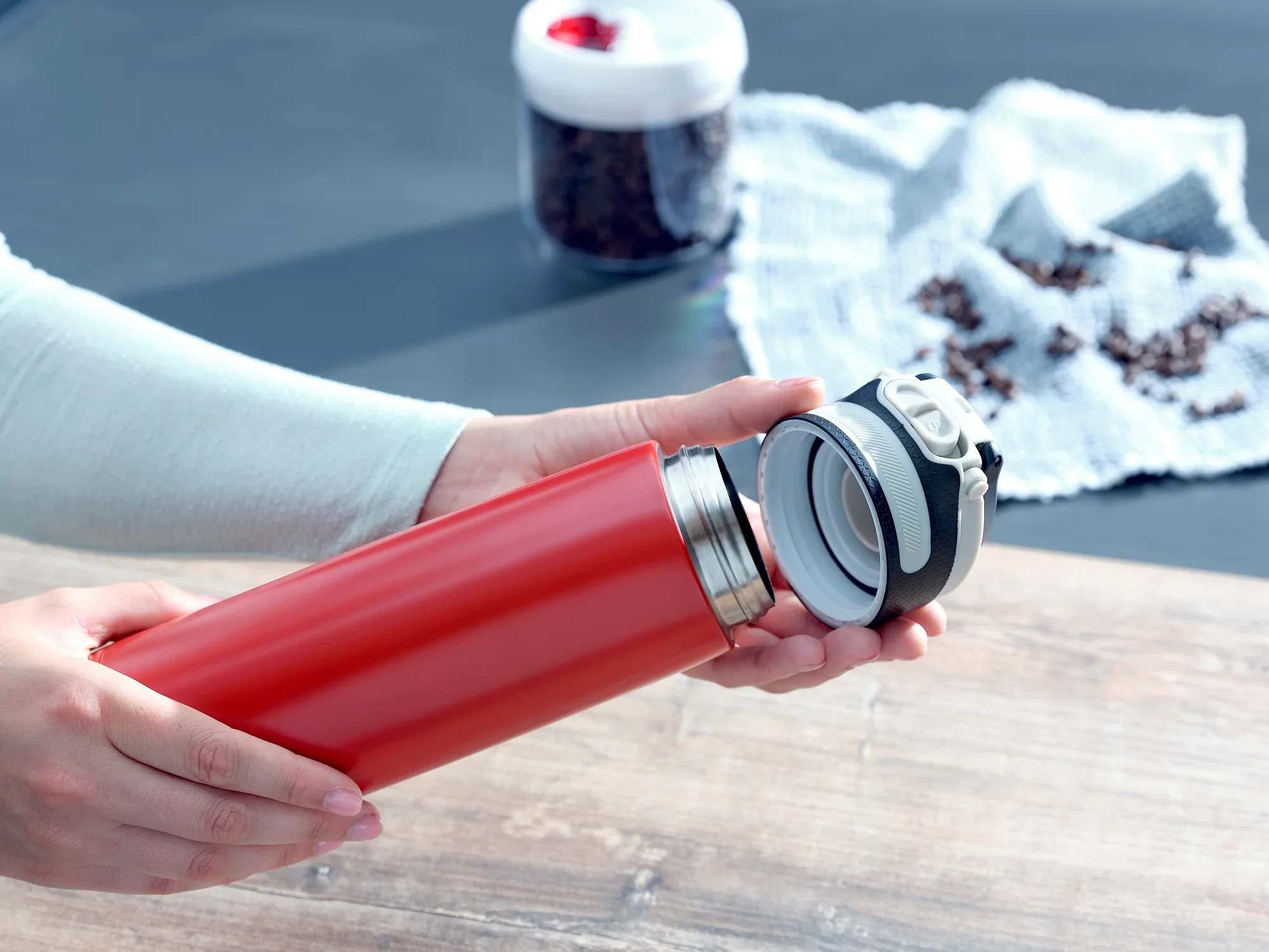 Osoba otwiera czerwony izolowany kubek Flip o pojemności 600 ml. W tle widać ziarna kawy i szklany pojemnik. Idealny do podróży.