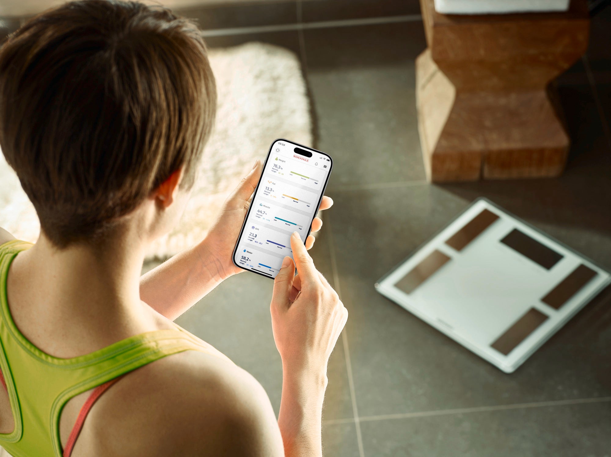 Eine Person betrachtet ein Smartphone, das die App der Personenwaage digital Shape Sense Connect 100 zeigt. Die Waage liegt auf dem Boden in einem modernen Raum.