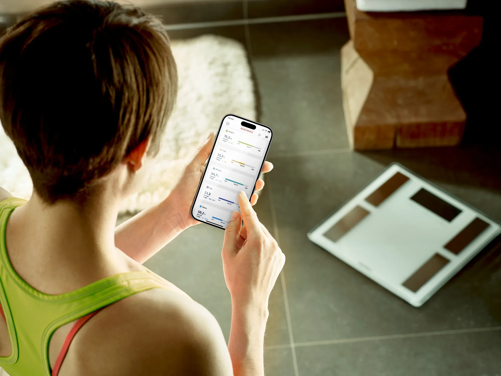 Een persoon kijkt naar een smartphone met daarop de app voor de Shape Sense Connect 100 digitale badkamerweegschaal. De weegschaal ligt op de vloer in een moderne kamer.