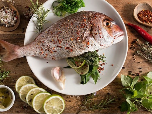 Ein gewuerzter Fisch liegt auf einem weissen Teller, umgeben von Limettenscheiben, Kraeutern, Knoblauch und Chili auf Holzuntergrund.