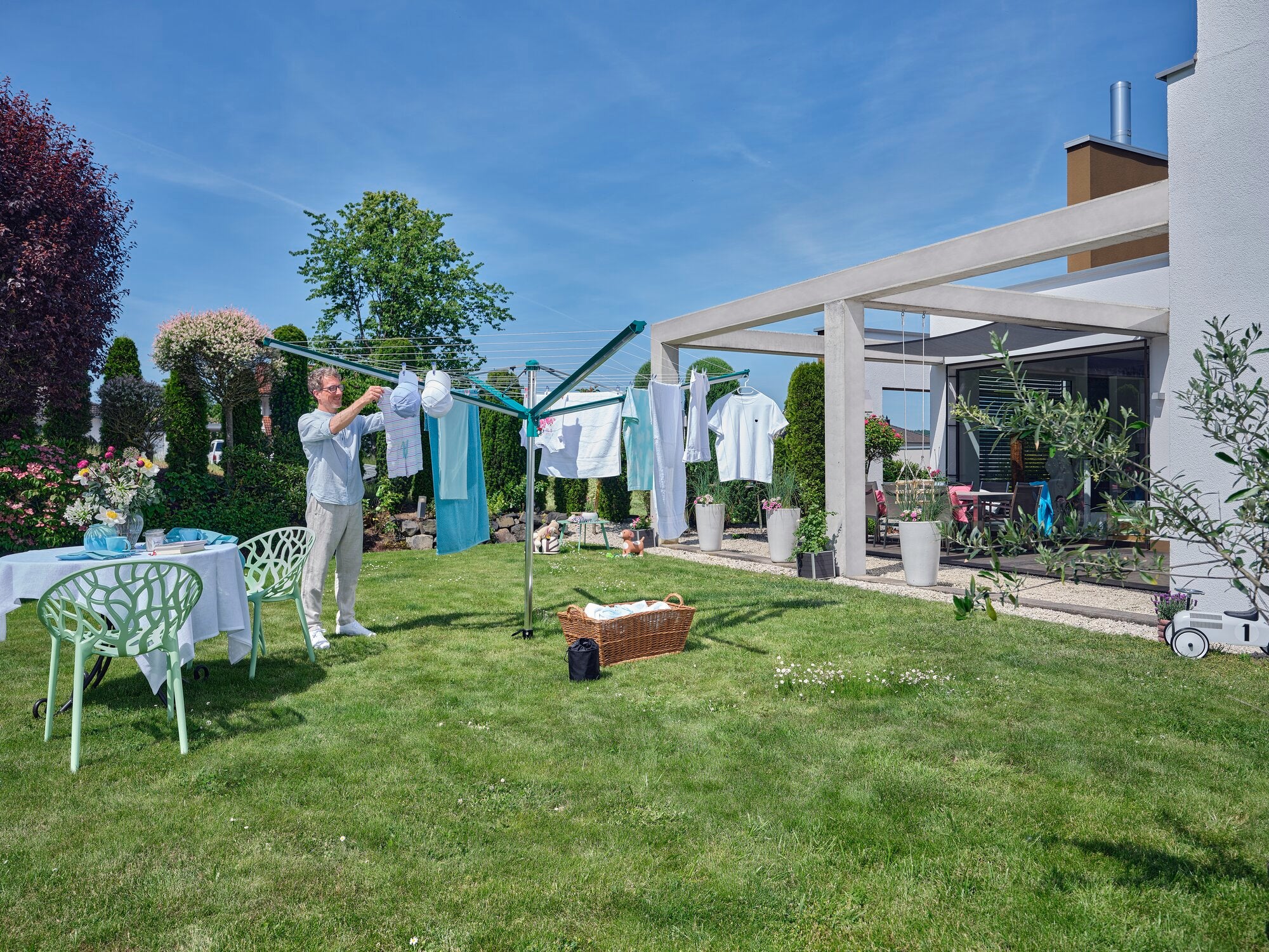 Una persona stende il bucato sull'asciugatrice Linomatic 500 Deluxe in giardino. L'ambiente è verde e soleggiato, con un tavolo e delle sedie sullo sfondo.