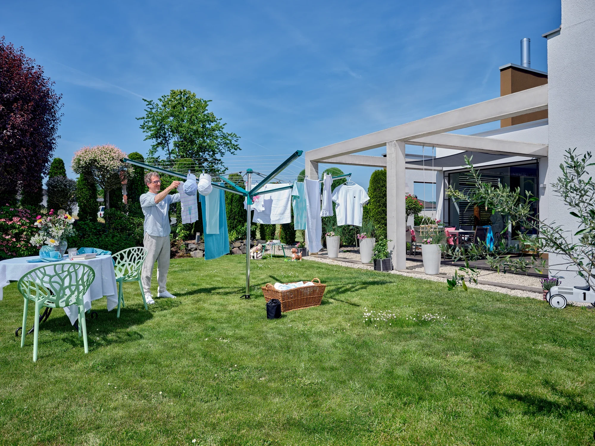Jedna osoba rozwiesza pranie na suszarce obrotowej Linomatic 500 Deluxe w ogrodzie. Otoczenie jest zielone i słoneczne, a w tle widać stół i krzesła.