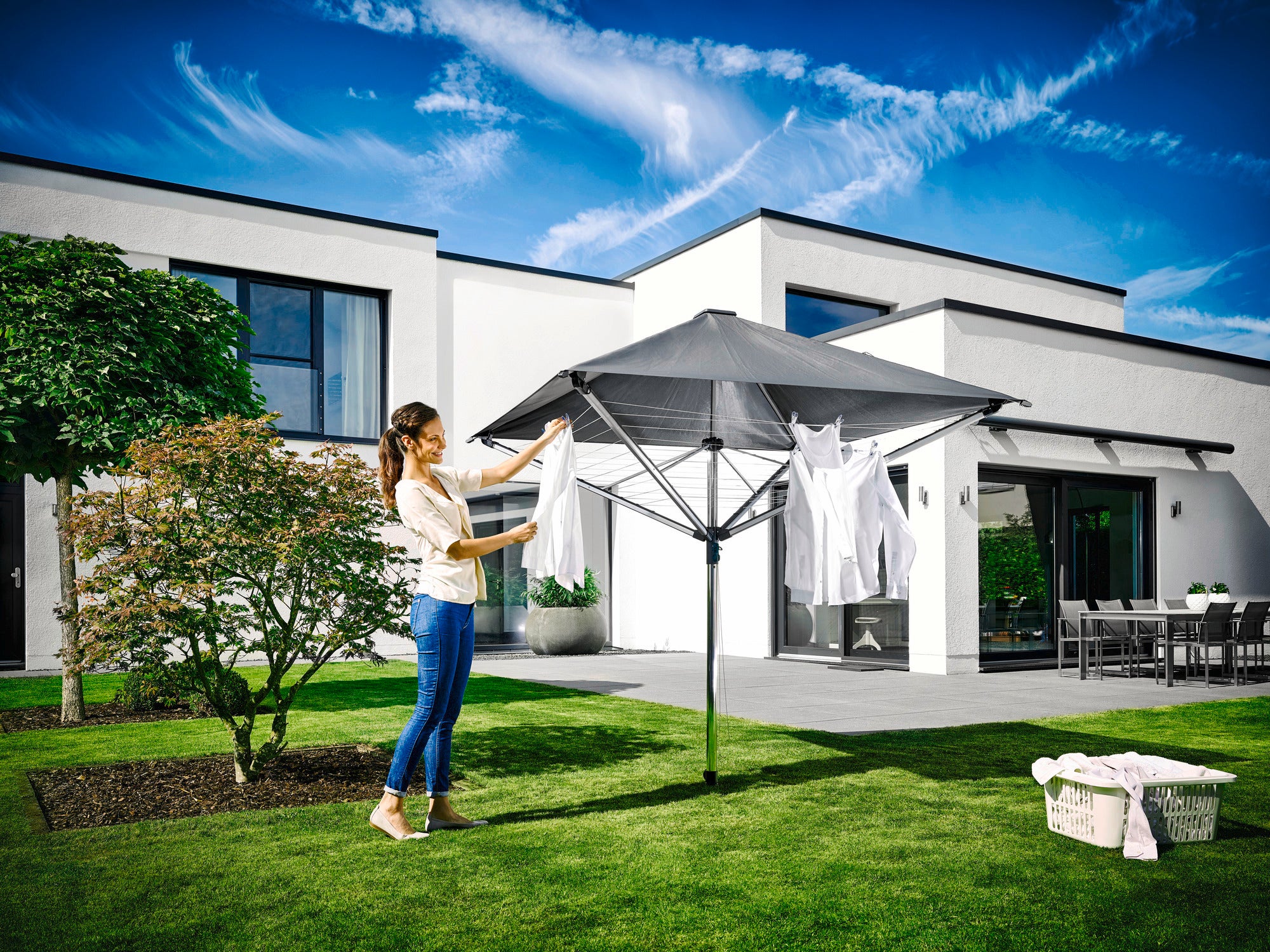 Een vrouw hangt wasgoed op de LinoProtect 400 droogmolen in een moderne tuin. De droogmolen biedt bescherming tegen regen en zon en past tegelijkertijd stijlvol in de omgeving.