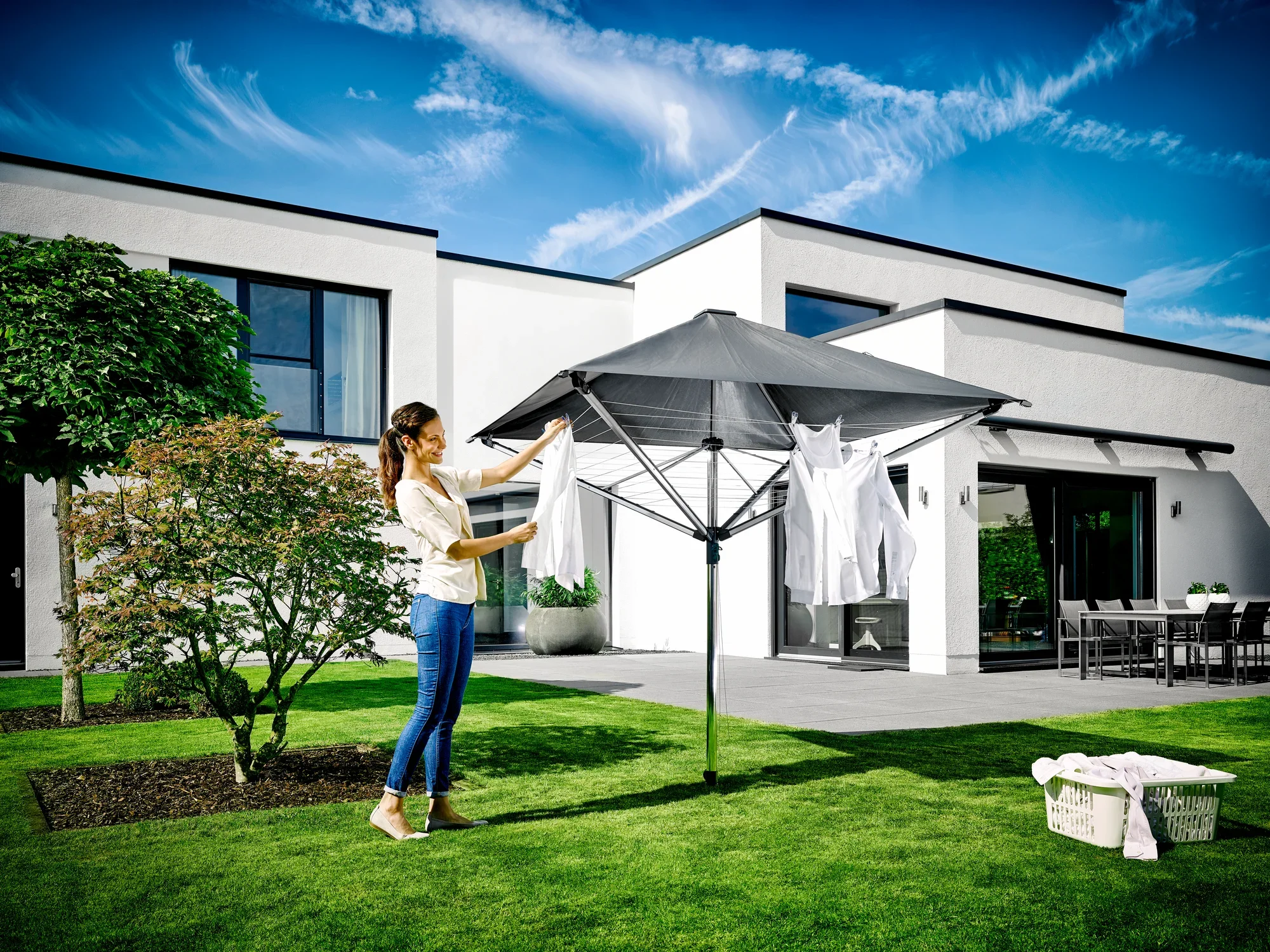 Une femme suspend du linge sur le séchoir à linge LinoProtect 400 dans un jardin moderne. Le séchoir à linge offre une protection contre la pluie et le soleil tout en s'intégrant avec élégance dans l'environnement.