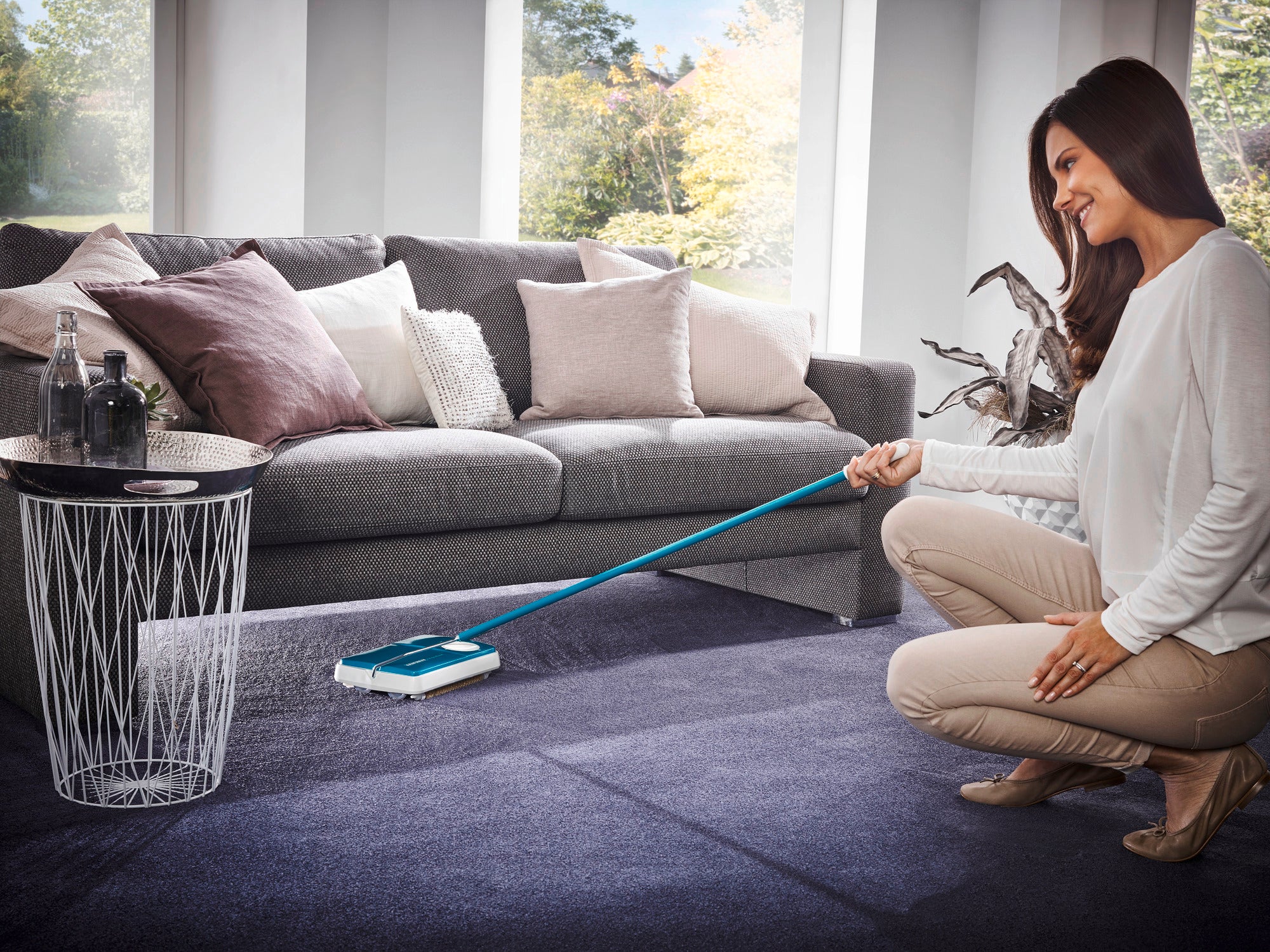 A woman uses the turquoise Regulus carpet sweeper to clean a dark blue carpet in a cosy living room with a grey sofa and cushions.