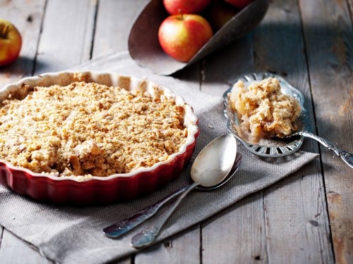 Runder Apfel-Crumble in roter Backform auf einem Holztisch, daneben liegen Loeffel und frische
