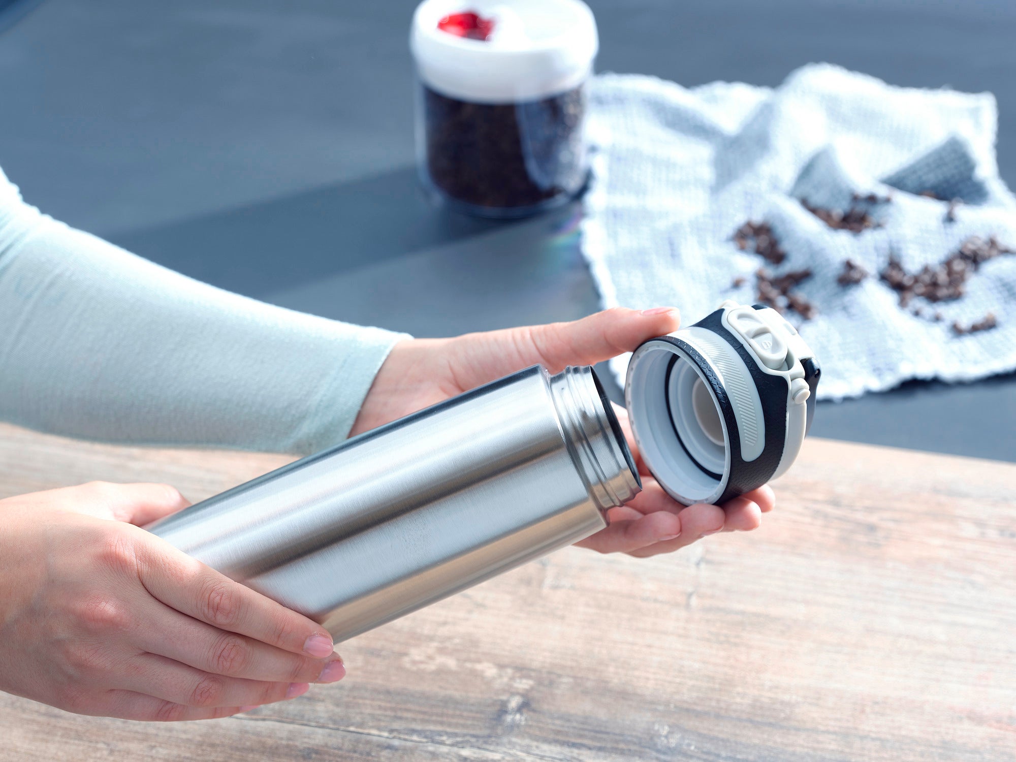 Isolierbecher Flip 600ml in Silber wird von einer Person geöffnet. Im Hintergrund sind Kaffeebohnen und ein Behälter zu sehen. Ideal für unterwegs, um Getränke warm zu halten.