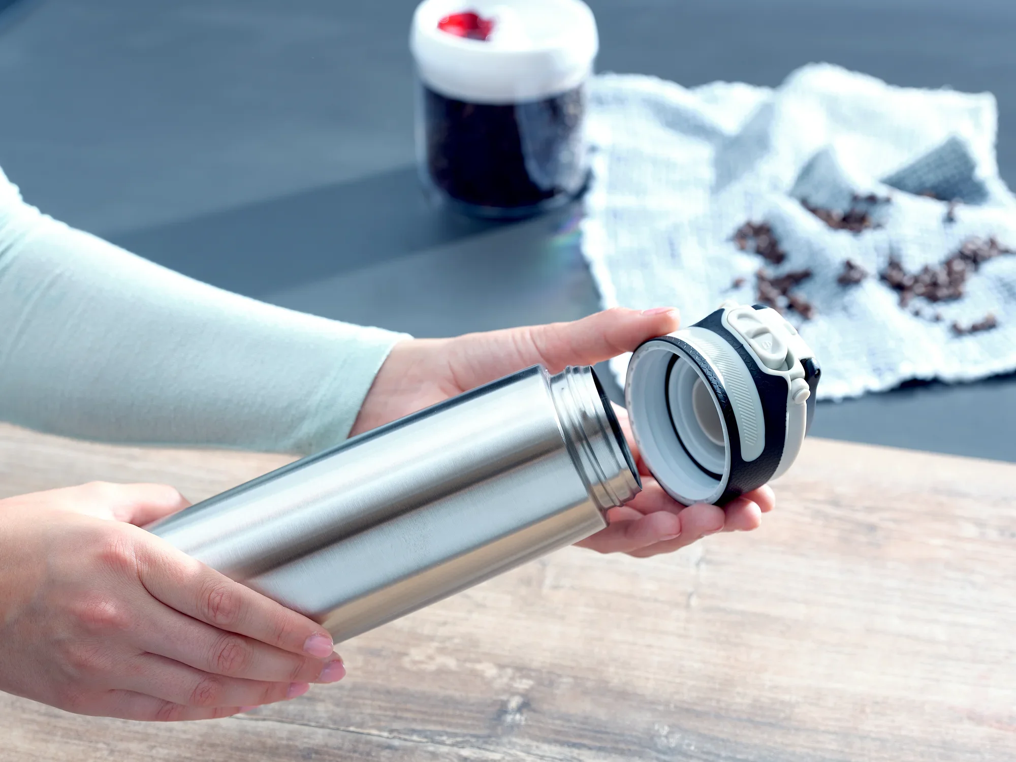 Izolowany kubek Flip 600ml w kolorze srebrnym otwierany przez osobę. W tle widoczne są ziarna kawy i pojemnik. Idealny do utrzymywania ciepła napojów podczas podróży.