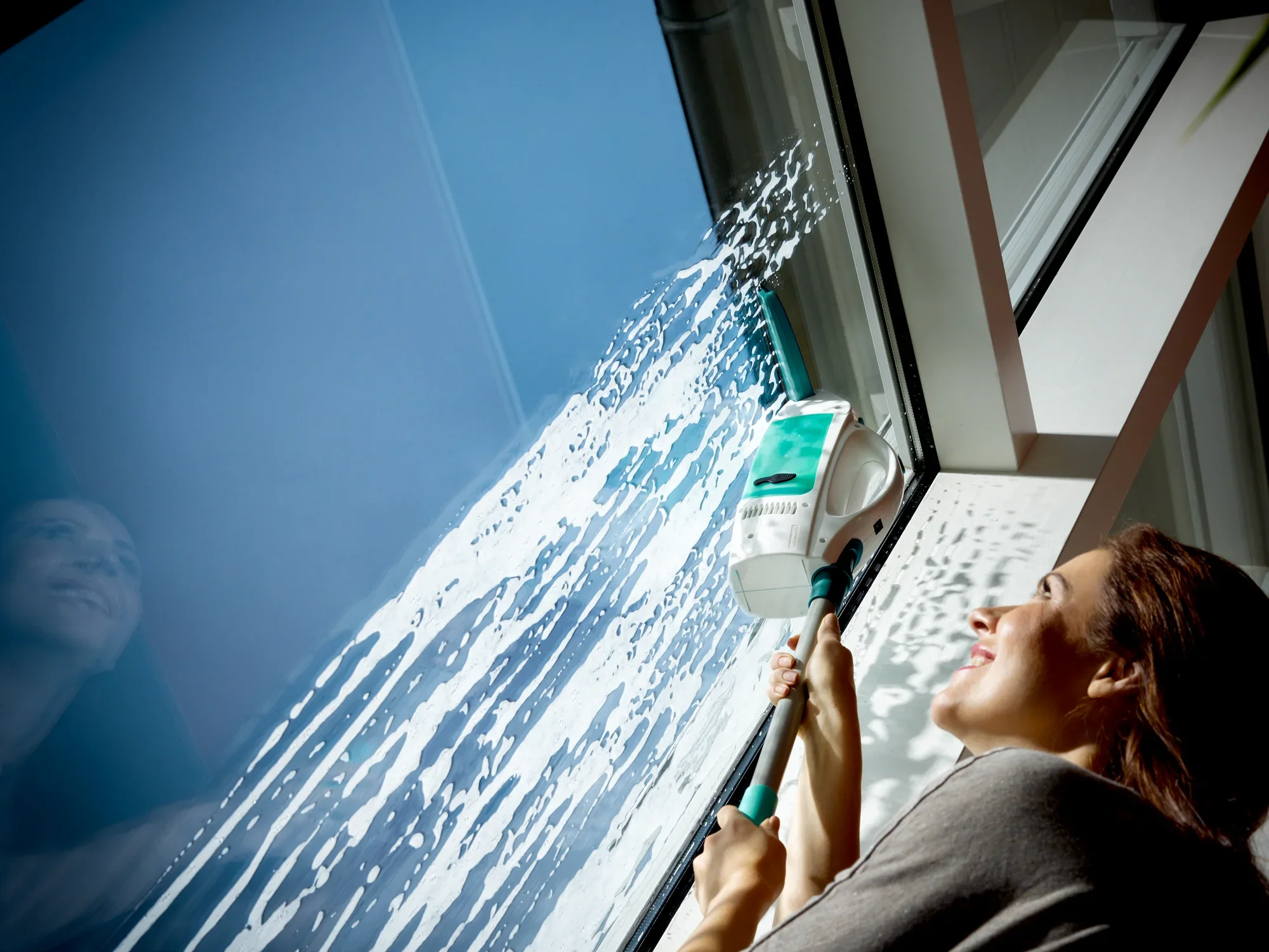 Kobieta myje duże okno za pomocą odkurzacza do okien Dry and Clean. Odkurzacz skutecznie usuwa wodę i brud, podczas gdy kobieta uśmiecha się, a słońce świeci do środka.