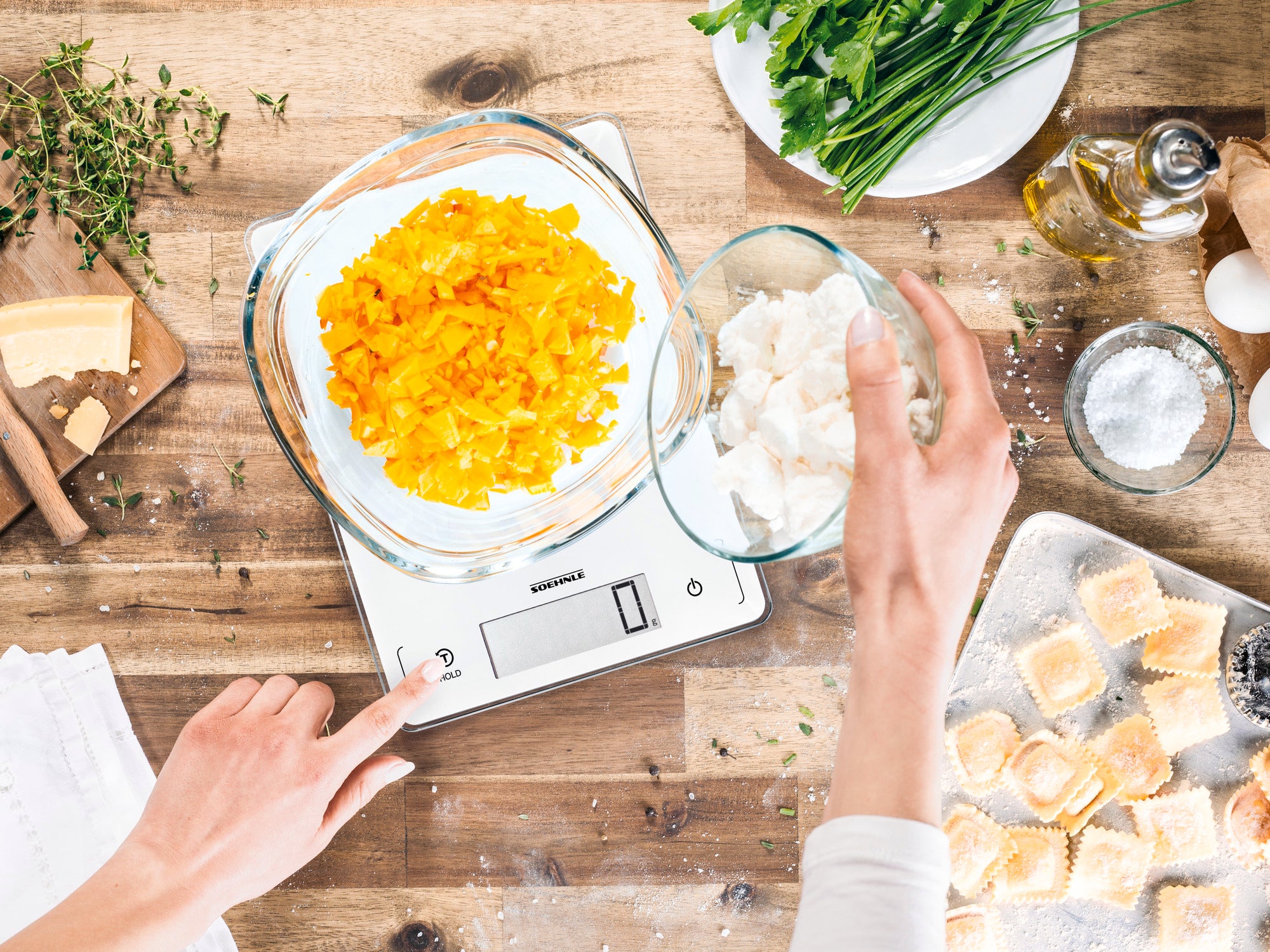 Eine digitale Küchenwaage zeigt eine Schüssel mit geschnittenen gelben Paprikaschoten und eine weitere Schüssel mit Käse. Eine Hand bedient die Waage, während Zutaten auf einem Holztisch verteilt sind.