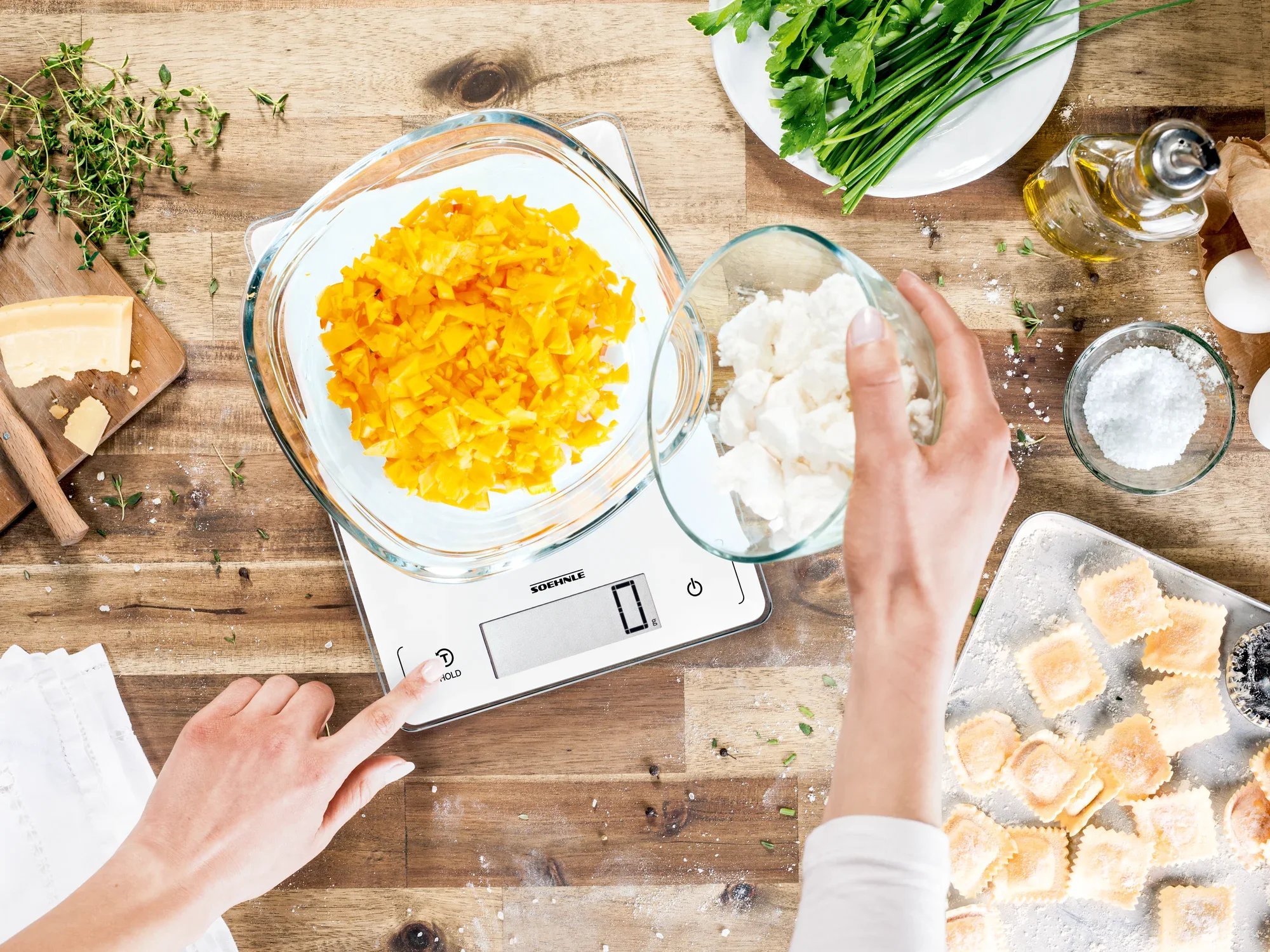 Eine digitale Küchenwaage zeigt eine Schüssel mit geschnittenen gelben Paprikaschoten und eine weitere Schüssel mit Käse. Eine Hand bedient die Waage, während Zutaten auf einem Holztisch verteilt sind.