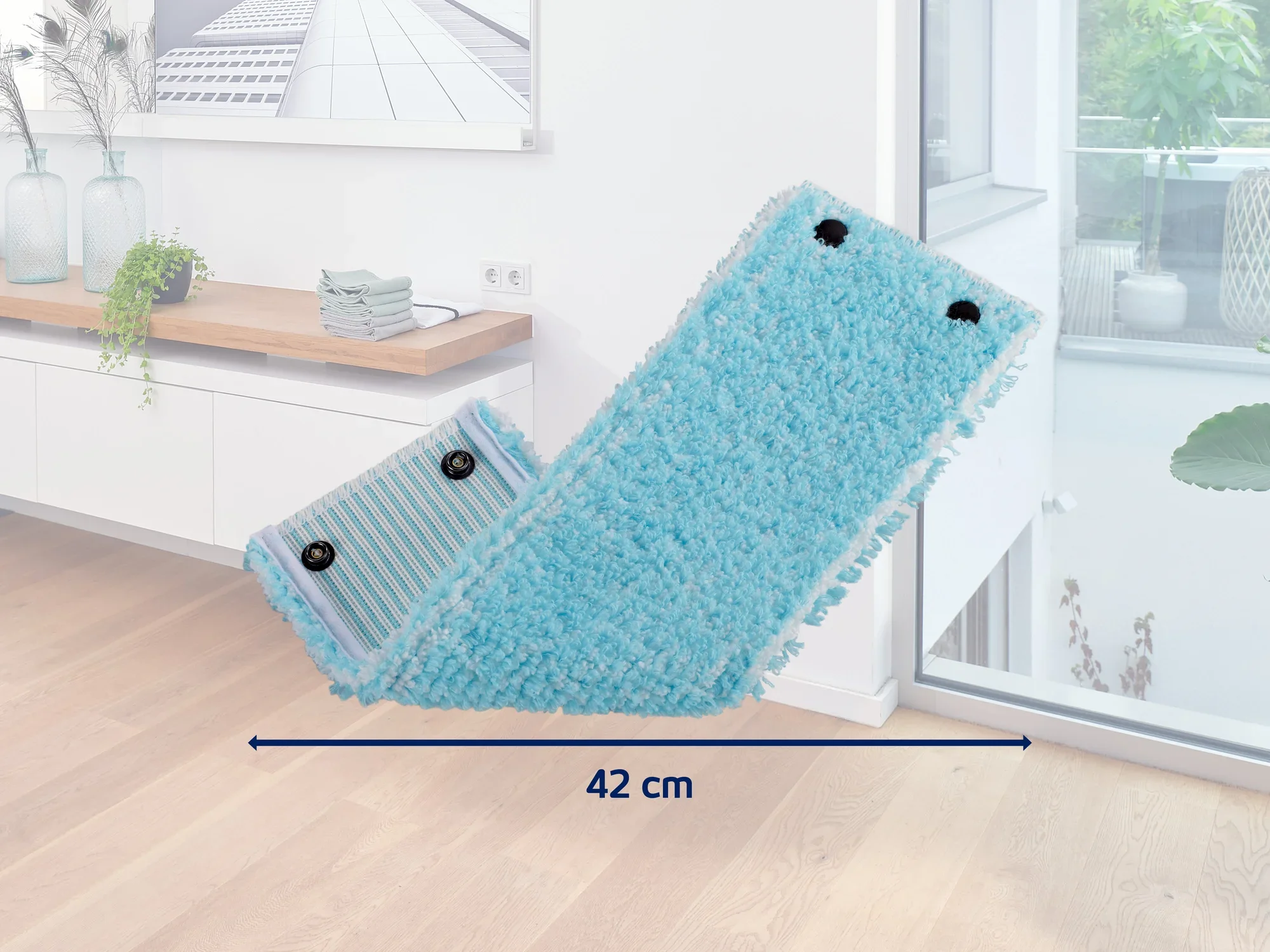 Un accesorio de mopa azul con un ancho de 42 cm se encuentra en una habitación moderna y luminosa con suelo de madera. Al fondo se ven plantas y decoraciones en una estantería.