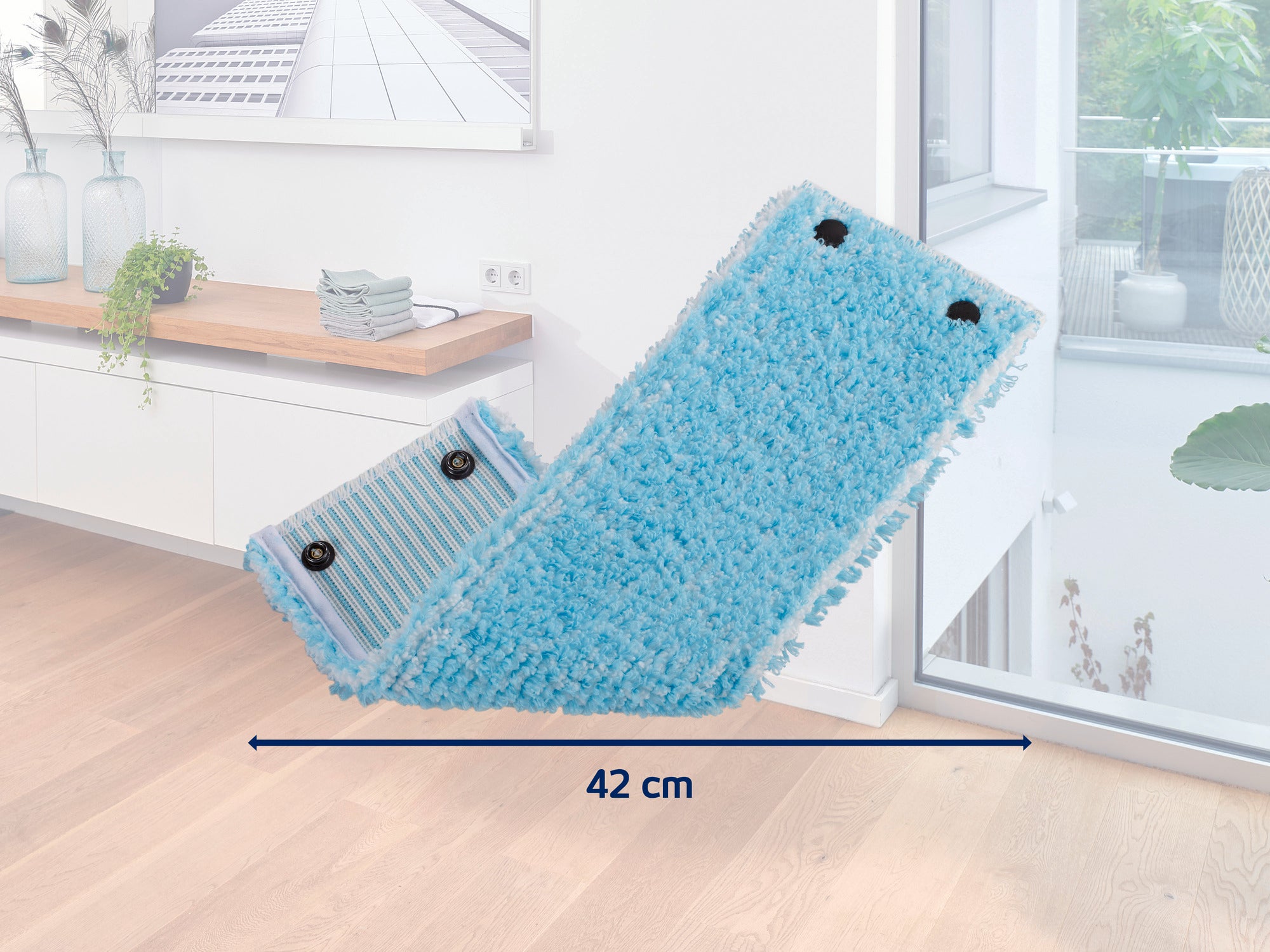 Ein blauer Wischmopp-Aufsatz mit einer Breite von 42 cm liegt in einem modernen, hellen Raum mit Holzboden. Im Hintergrund sind Pflanzen und Dekorationen auf einem Regal zu sehen.