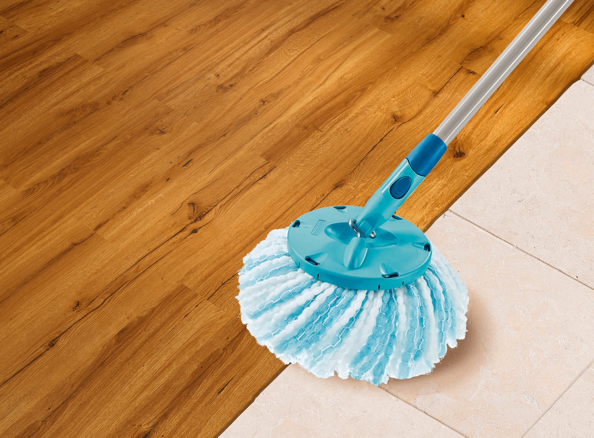 Ein CLEAN TWIST Disc Mop microduo Ersatzkopf wischt über einen Boden, der zur Hälfte aus Holz und zur Hälfte aus Fliesen besteht. Der Mop hat einen blauen Griff und weiße Fasern.