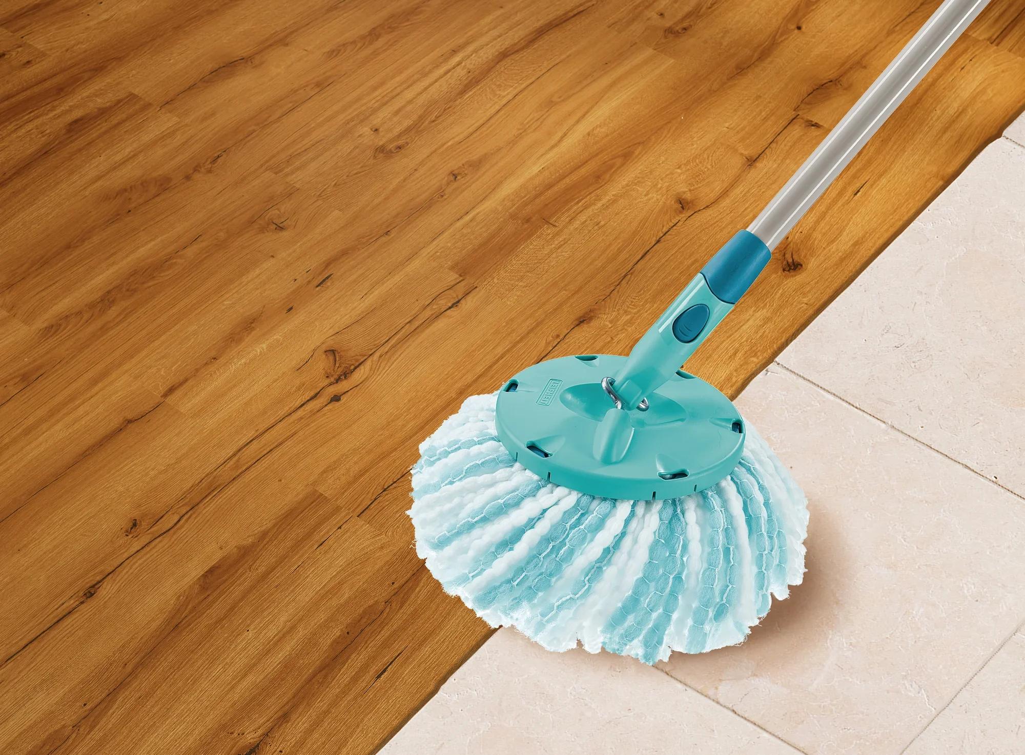 Ein CLEAN TWIST Disc Mop microduo Ersatzkopf wischt über einen Boden, der zur Hälfte aus Holz und zur Hälfte aus Fliesen besteht. Der Mop hat einen blauen Griff und weiße Fasern.