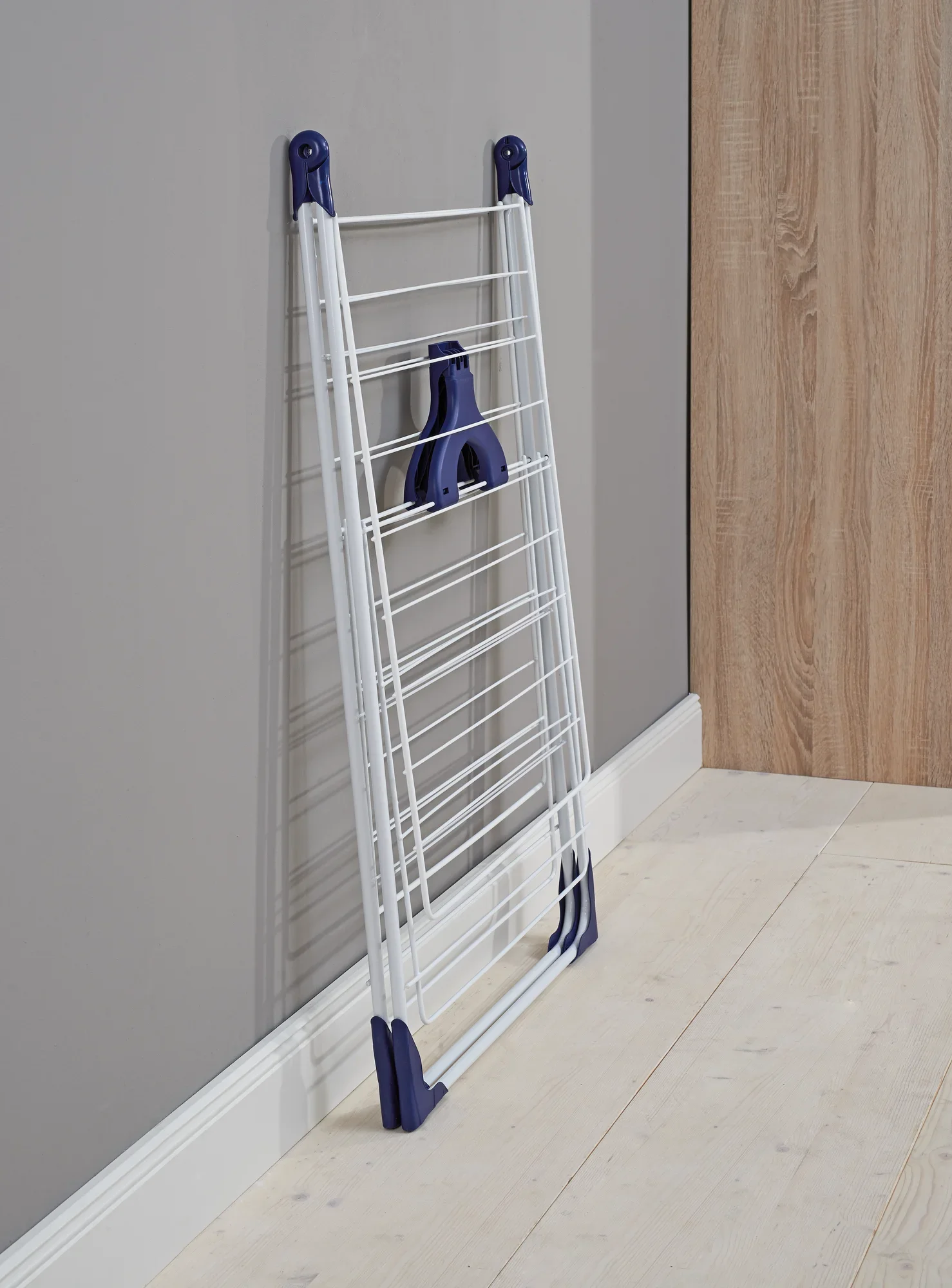 Suszarka wolnostojąca Classic 200 Flex: Koń ze złożonymi ubraniami opiera się o szarą ścianę. Ma niebieskie akcenty i jest idealna do małych przestrzeni.