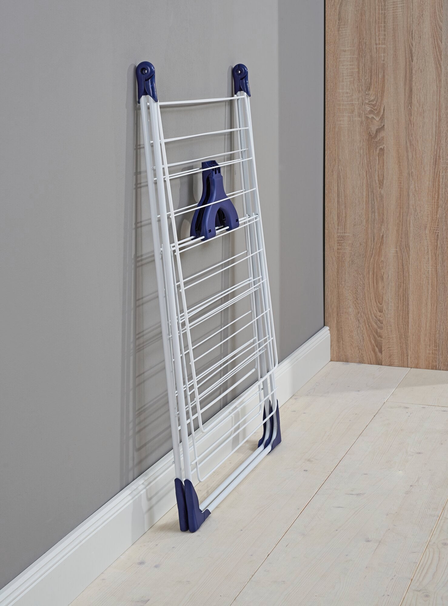 Classic 200 Flex vrijstaande droger: Een opgevouwen droogrek leunt tegen een grijze muur. Hij heeft blauwe accenten en is ideaal voor kleine ruimtes.