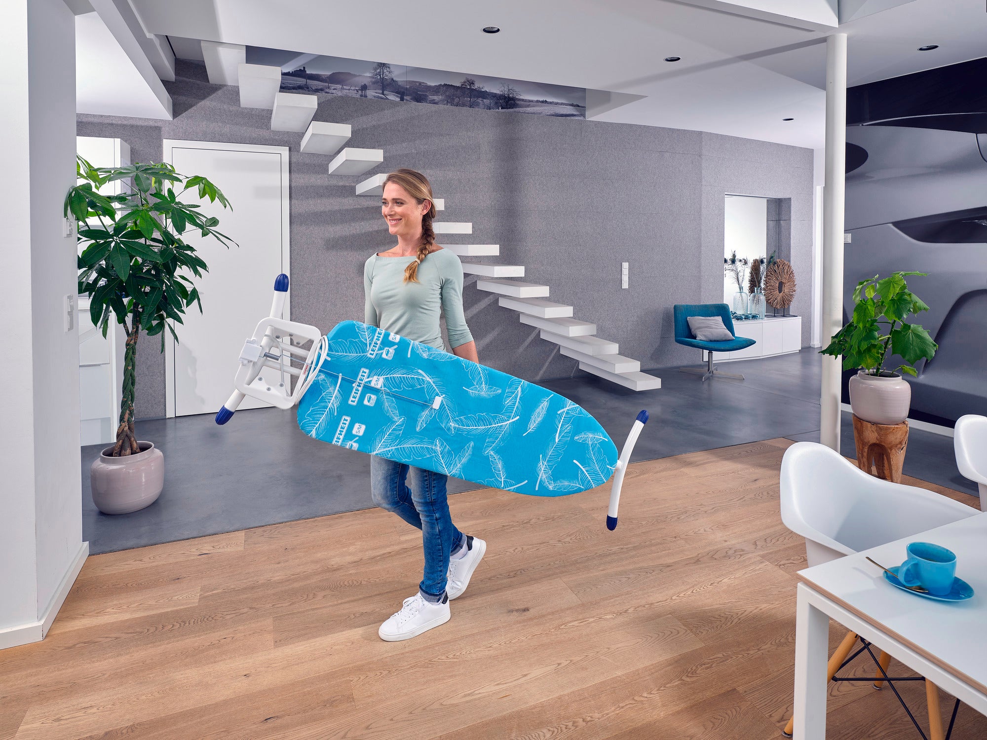 Eine Frau trägt den Bügeltisch Air Board M Solid Plus durch ein modernes Wohnzimmer. Der Tisch ist leicht und handlich, ideal für den täglichen Gebrauch.
