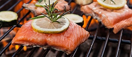 Auf einem heissen Grill liegen Lachsfilets mit Zitronenscheiben und Rosmarinzweigen, daneben liegen Zucchinischeiben.