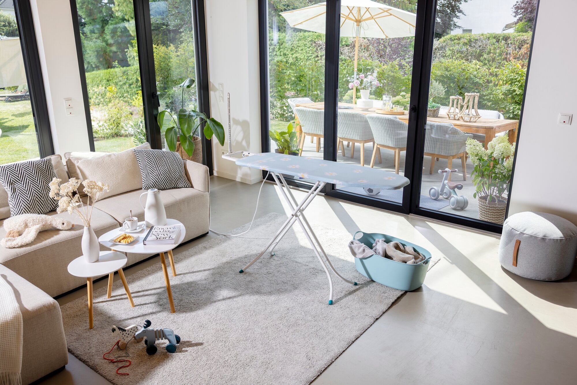 Bügelbrett Leifheit Classic Basic Plus in einem modernen Wohnzimmer mit großen Fenstern. Es steht auf einem Teppich neben einem Tisch mit Dekorationen und einem Spielzeug.